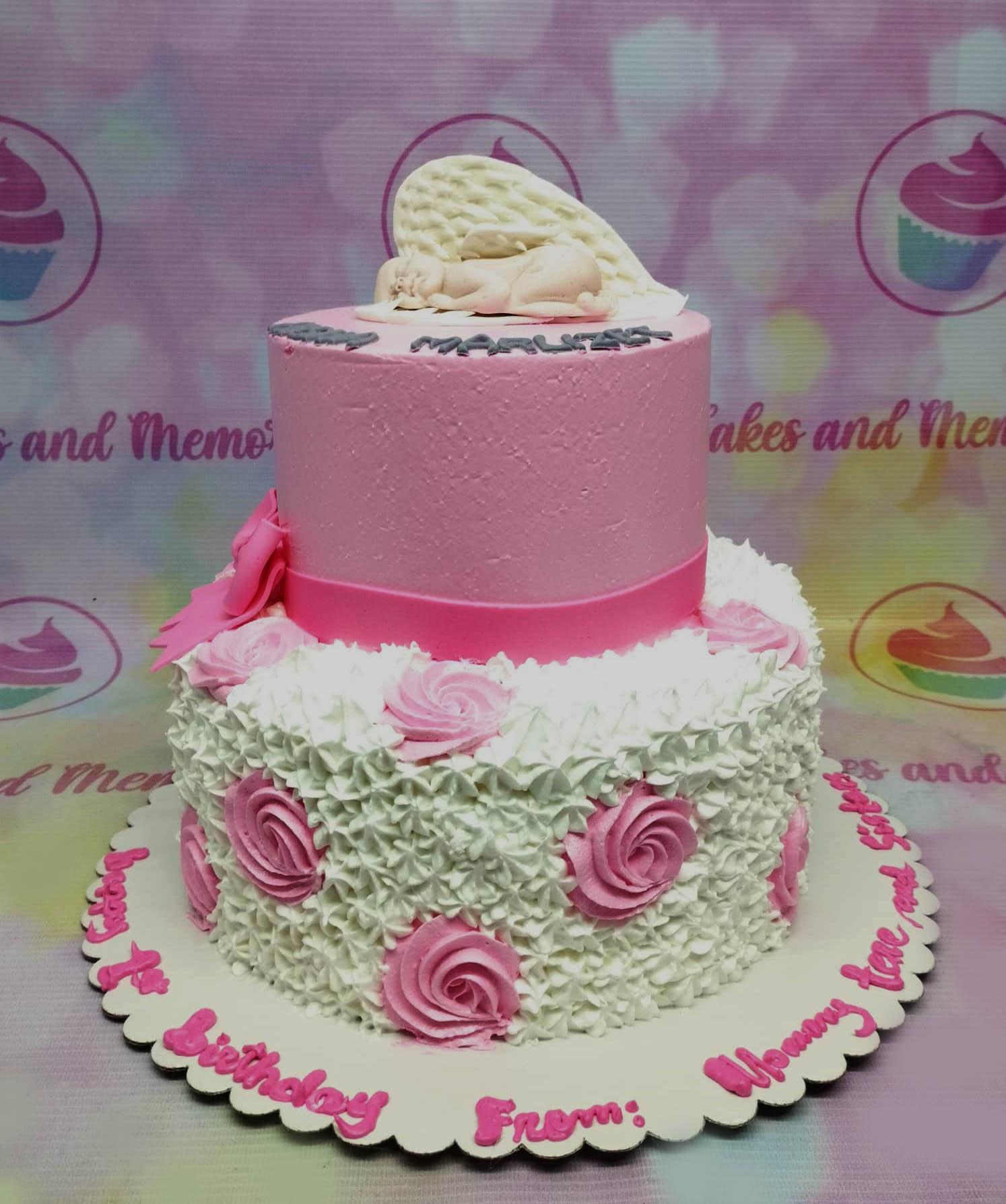 Two-tier pink and white cake with a sleeping baby angel topper and pink icing rosettes on the bottom tier.