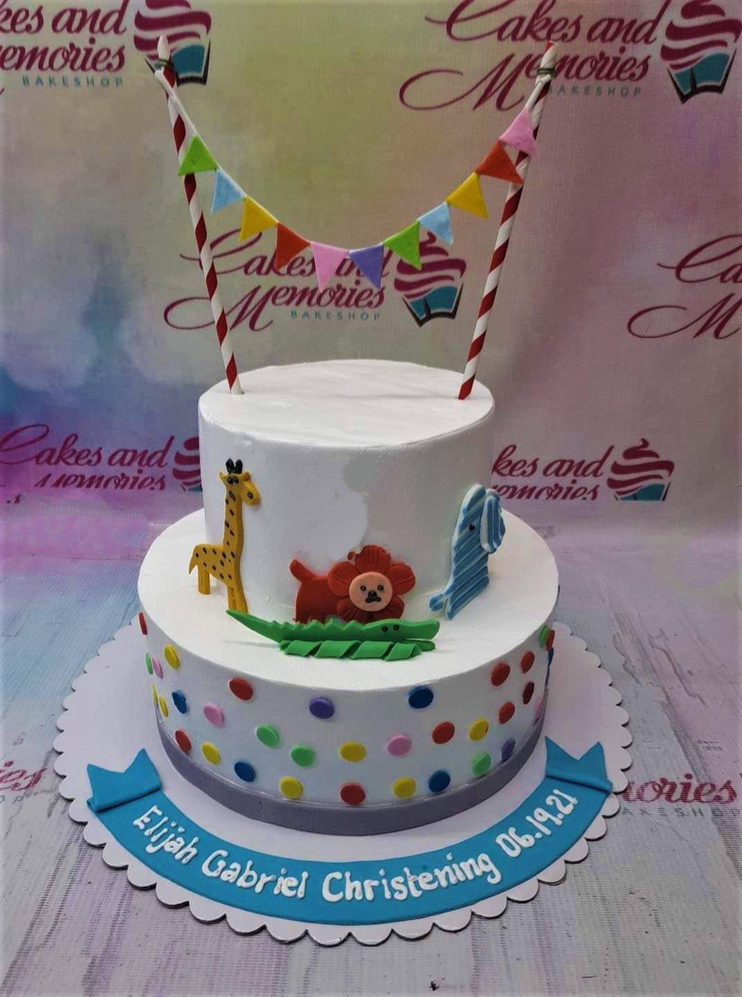 White 2-tier animal themed christening cake with fondant giraffe, lion, elephant, and crocodile figures and colorful polka dots.