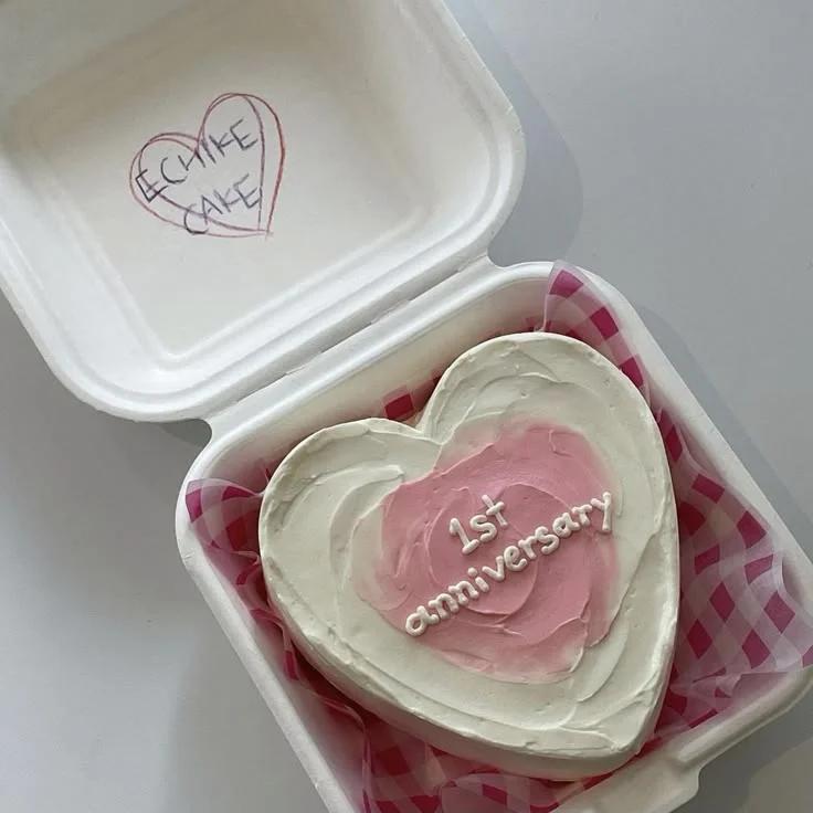 Heart-shaped white bento cake with a pink palette knife center and white piped 1st anniversary message in a white box.