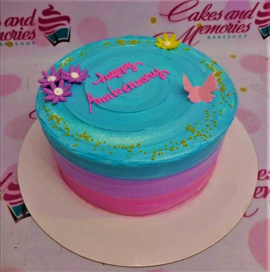 Round blue and pink gradient cake with purple flowers, a pink butterfly, and gold sprinkles for an anniversary celebration.