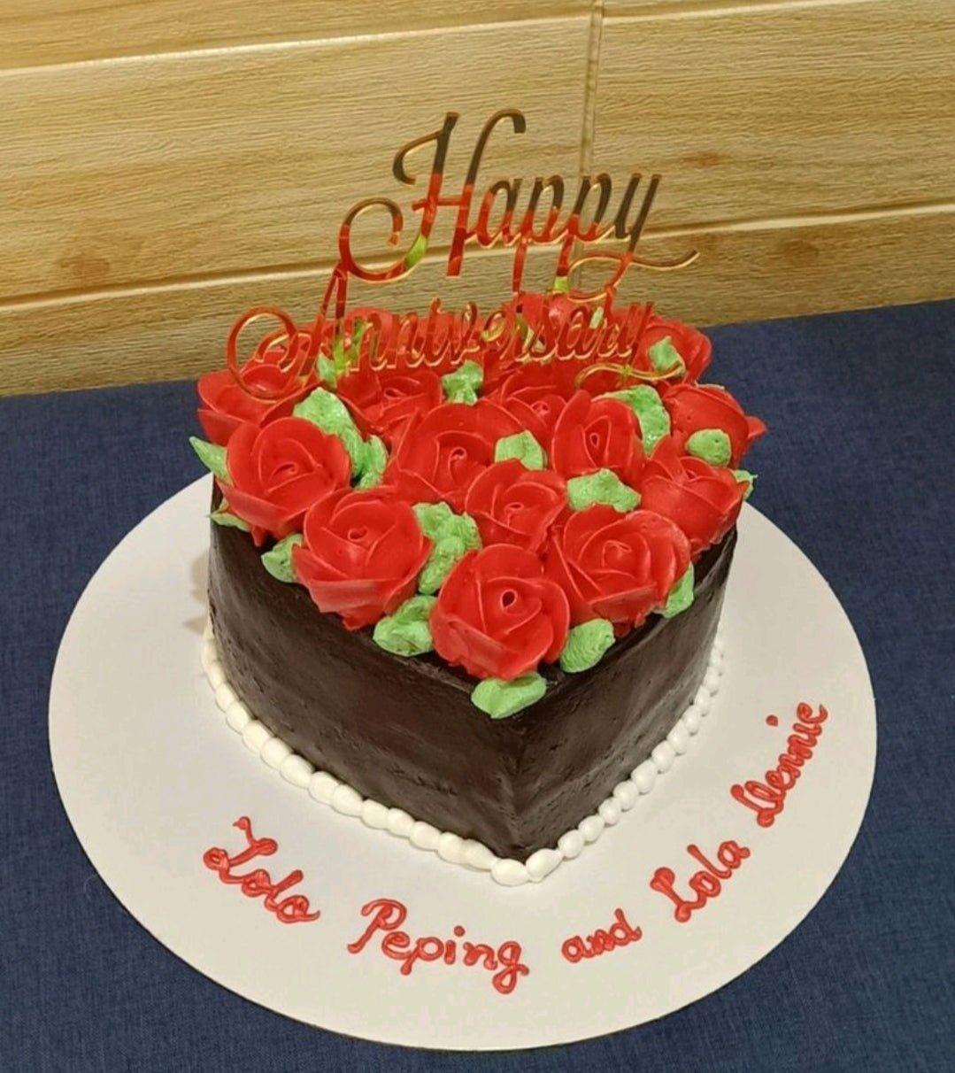 Heart-shaped chocolate cake with red icing roses and a gold Happy Anniversary topper on a white board with red piped message.