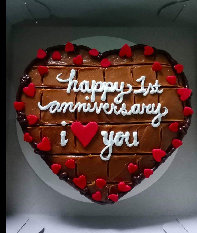 Heart-shaped chocolate cake with caramel icing, red fondant hearts, and white piped anniversary message.