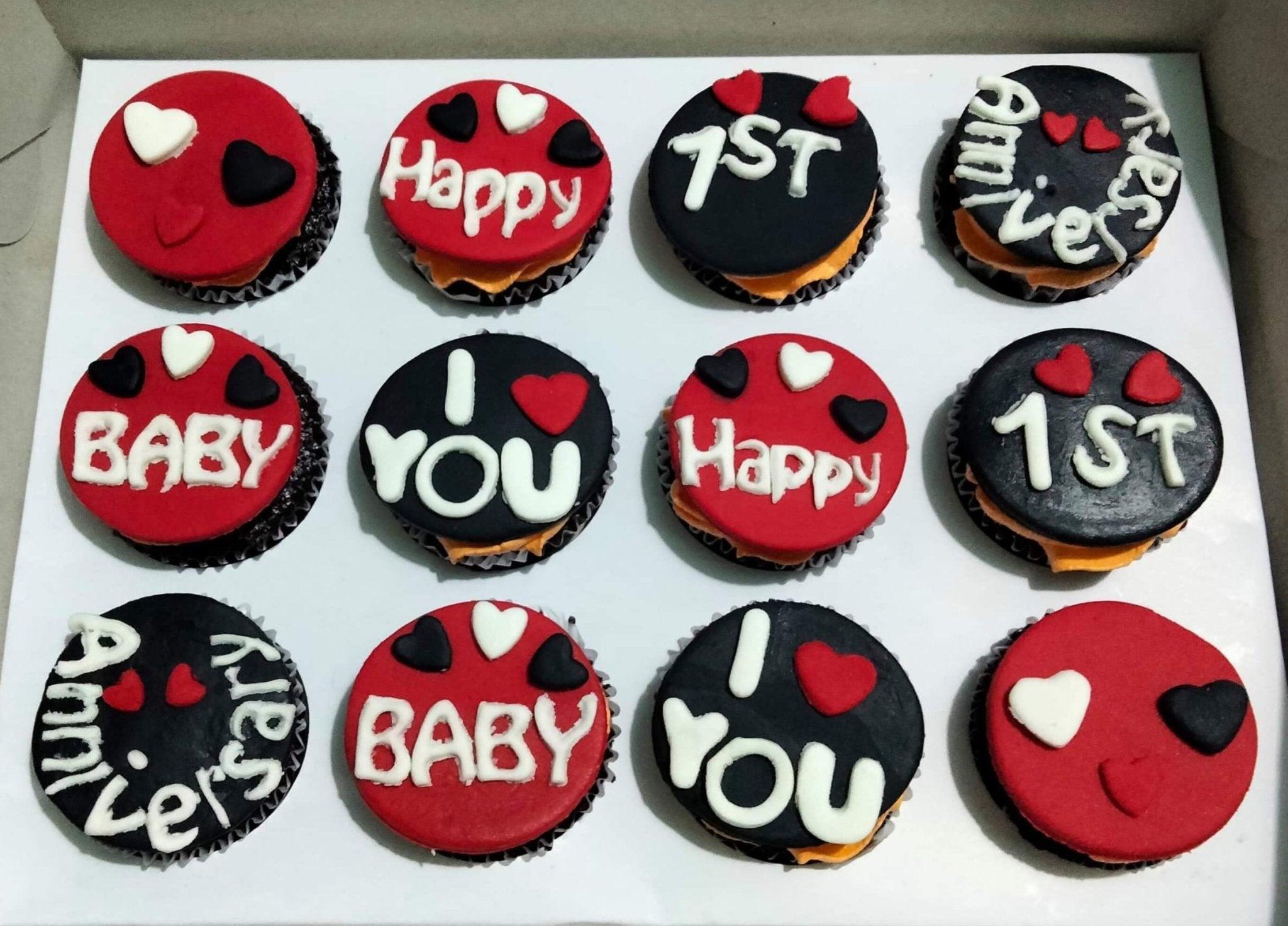 Twelve individual cupcakes with red and black fondant toppers featuring hearts and anniversary messages in white and red icing.