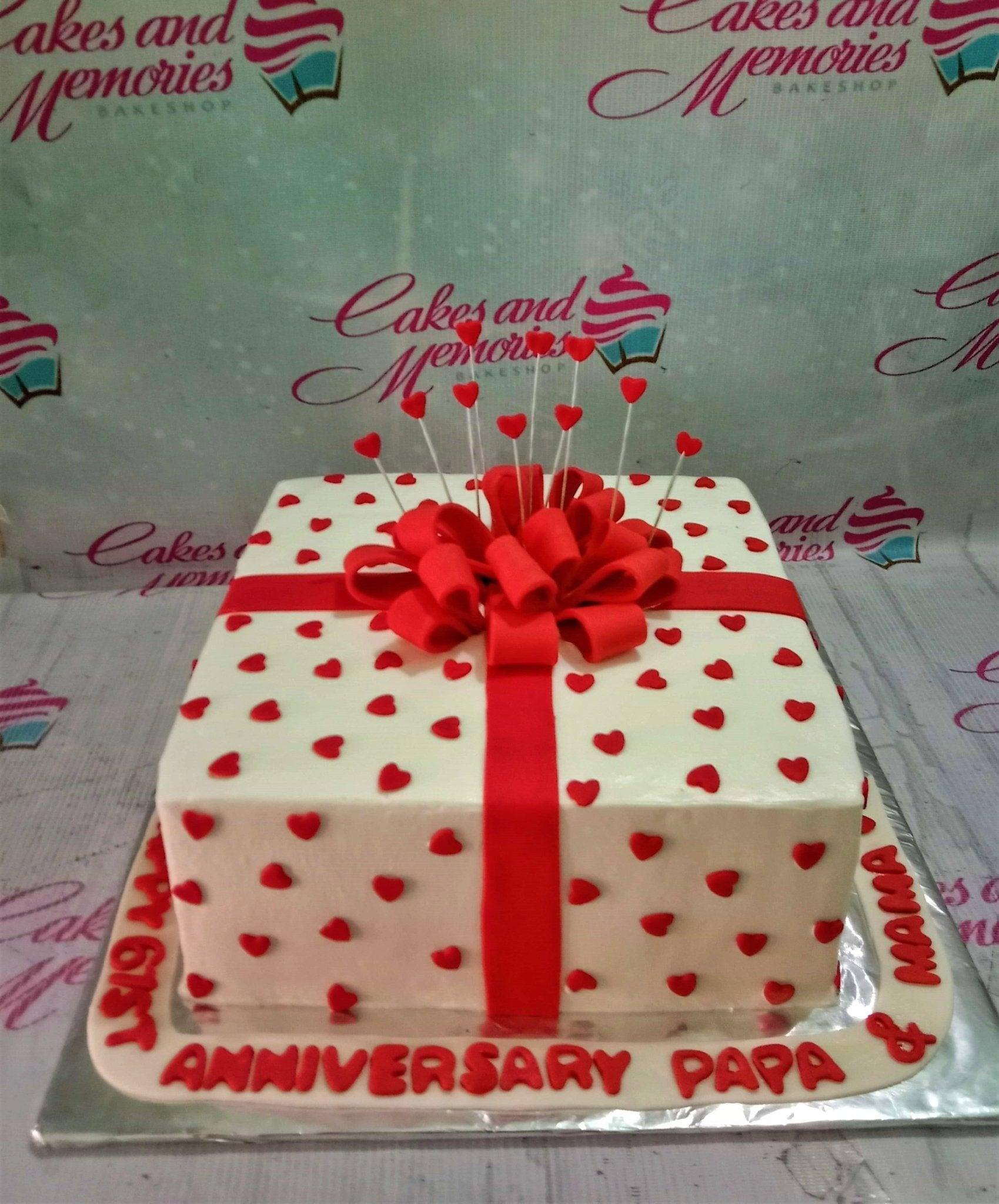 Square white cake designed like a gift box with a large red fondant bow and small red hearts all over.