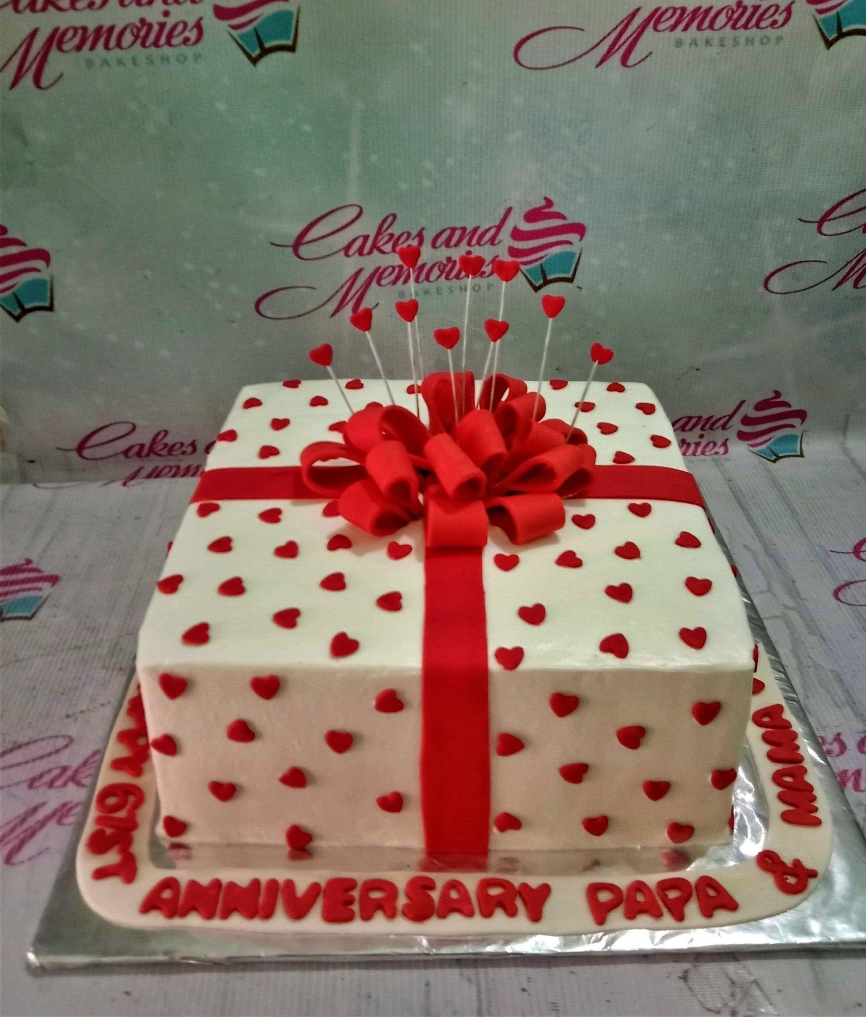 Square white gift box cake with red fondant hearts, a large red fondant bow, and heart toppers on sticks for a 61st anniversary.