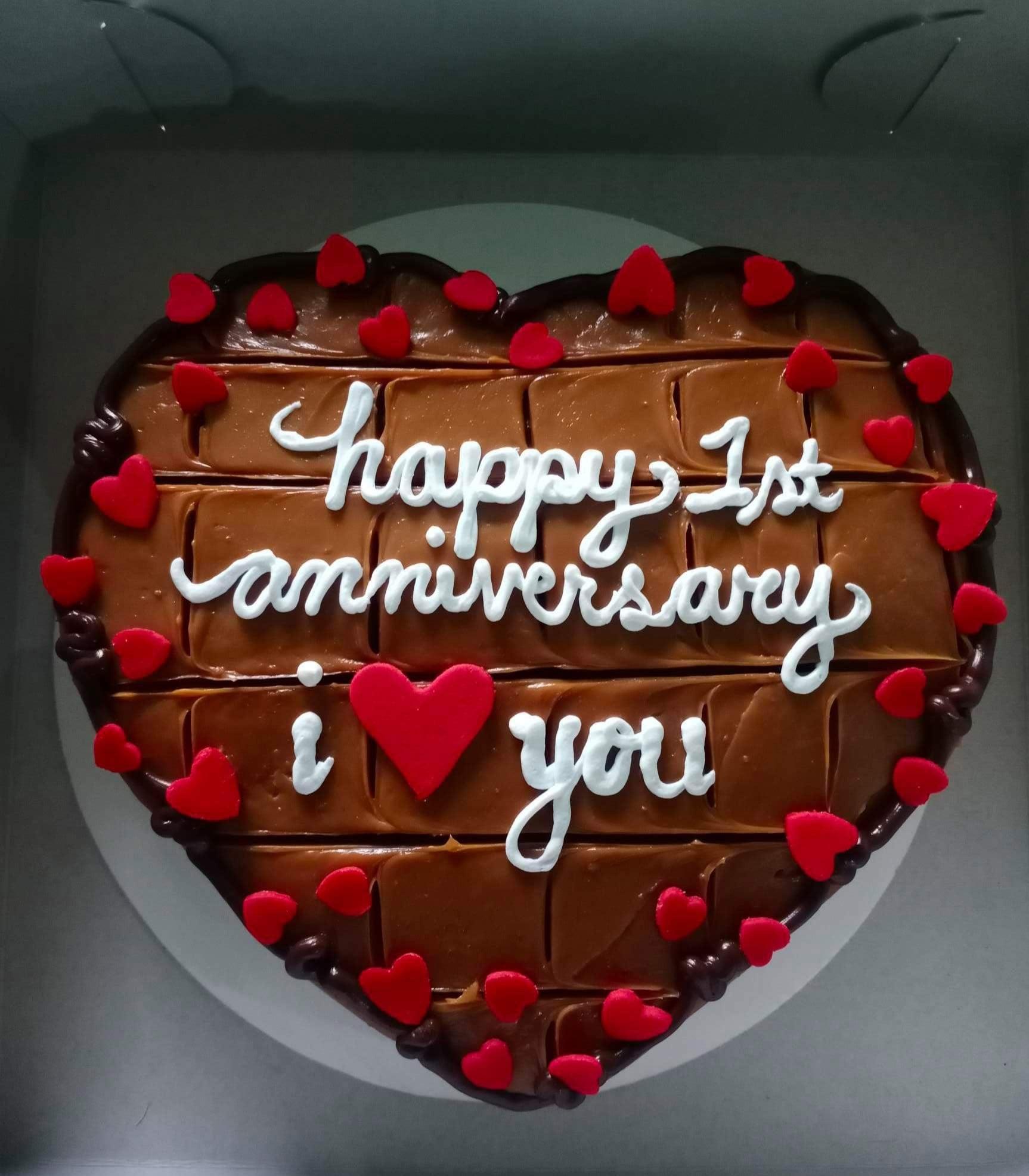 Heart-shaped chocolate cake with caramel icing, red fondant hearts, and white piped anniversary message.