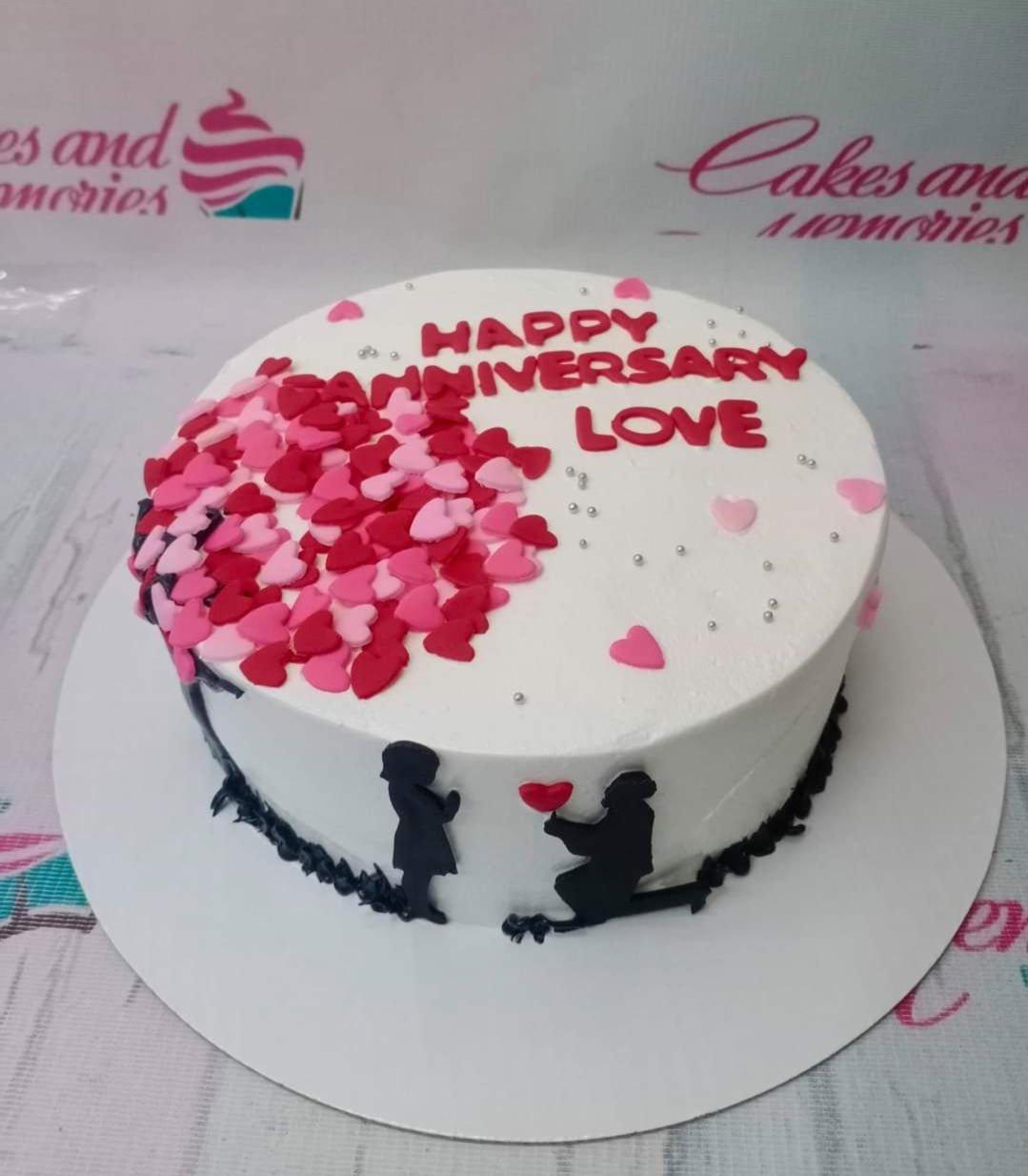 White anniversary cake with a red heart tree design, silver dragees, and black silhouettes of a couple proposing.