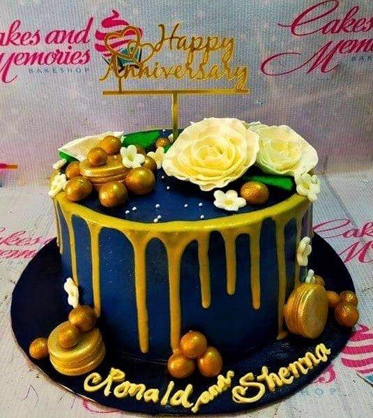 Navy blue anniversary cake with gold drip, white roses, gold macarons, and a gold Happy Anniversary topper.