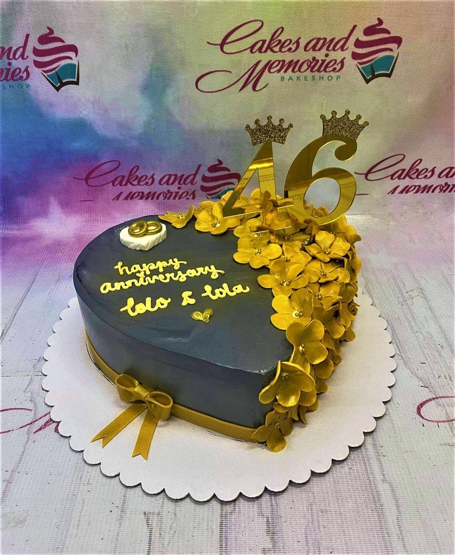 Heart-shaped navy blue anniversary cake with gold fondant flowers, gold number 46 toppers with crowns, and a gold ribbon bow.