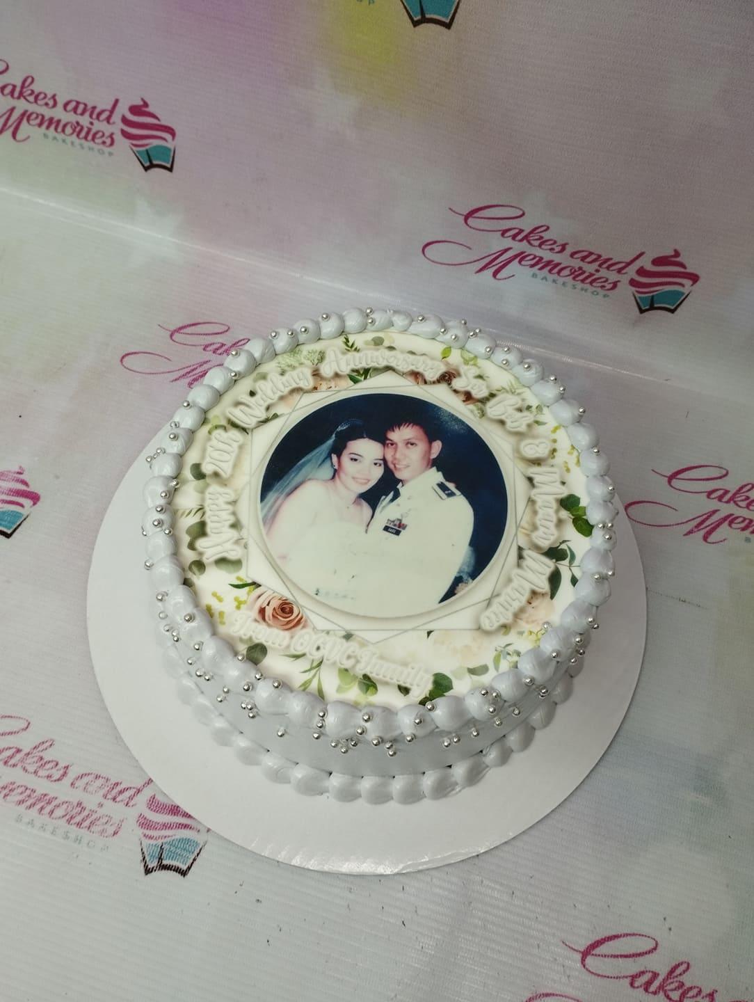 White 1-tier anniversary cake with a central edible photo print, floral designs, and silver dragees on soft icing swirls.