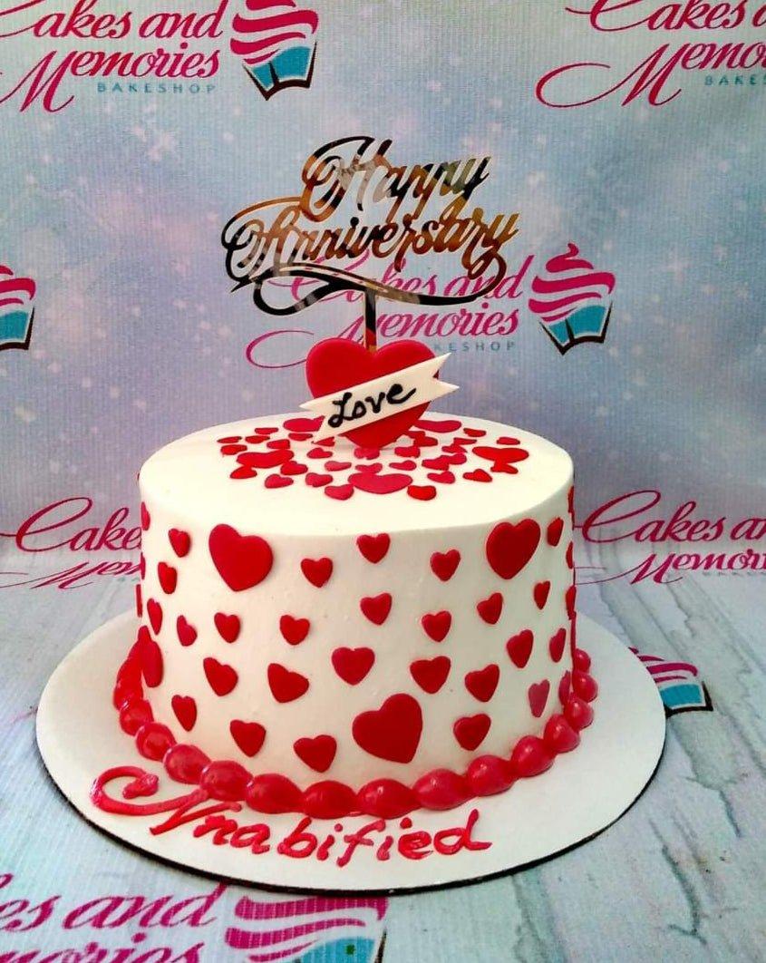 White anniversary cake decorated with numerous red fondant hearts and a gold Happy Anniversary topper.
