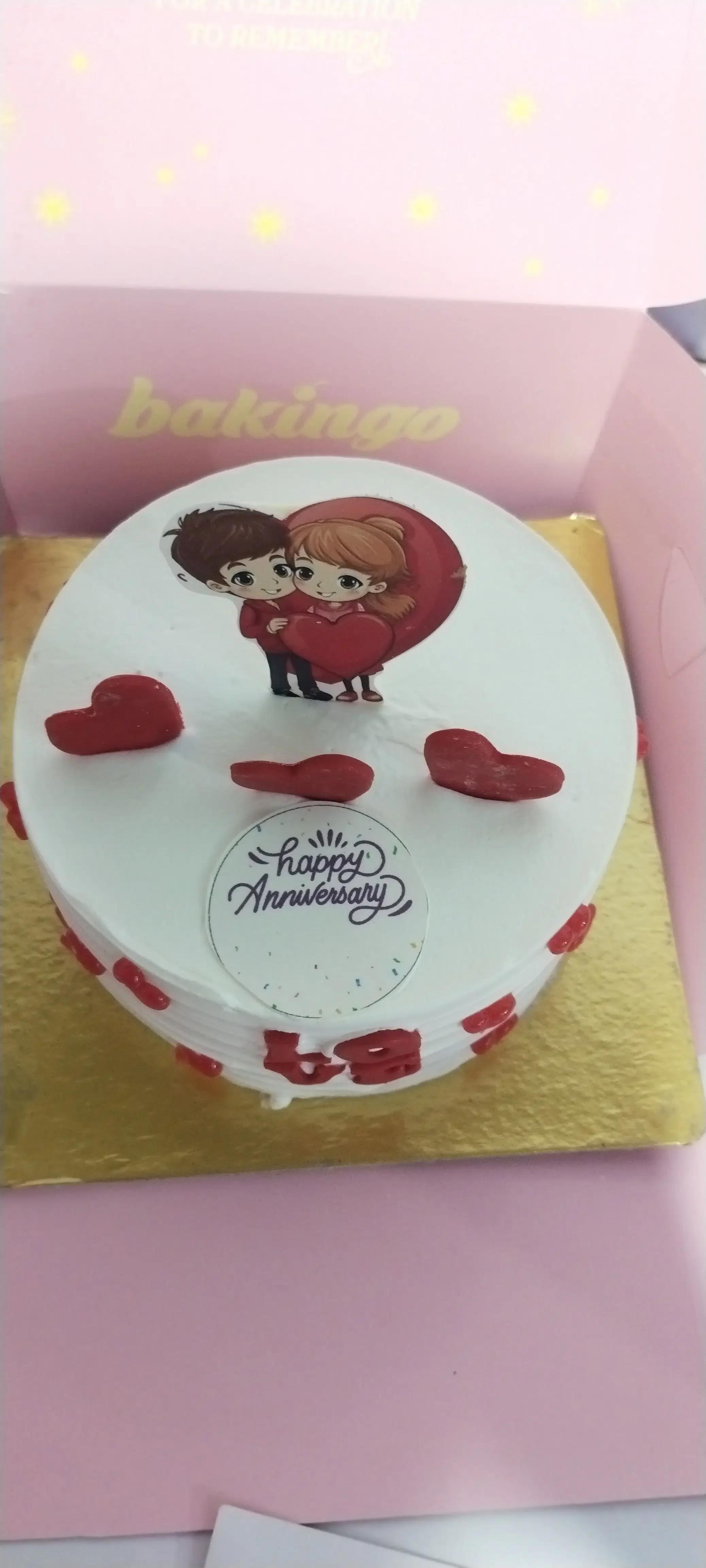 White round anniversary cake with a cartoon couple printout, red fondant hearts, and a Happy Anniversary message.