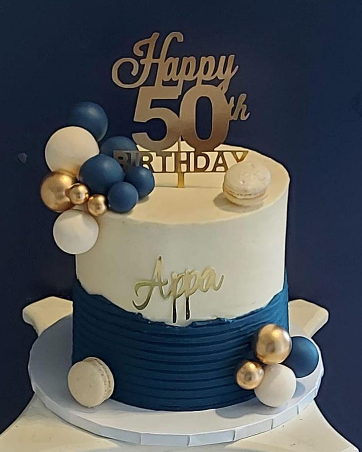 Elegant white and navy blue 50th birthday cake for Appa with gold accents, macarons, and decorative spheres.