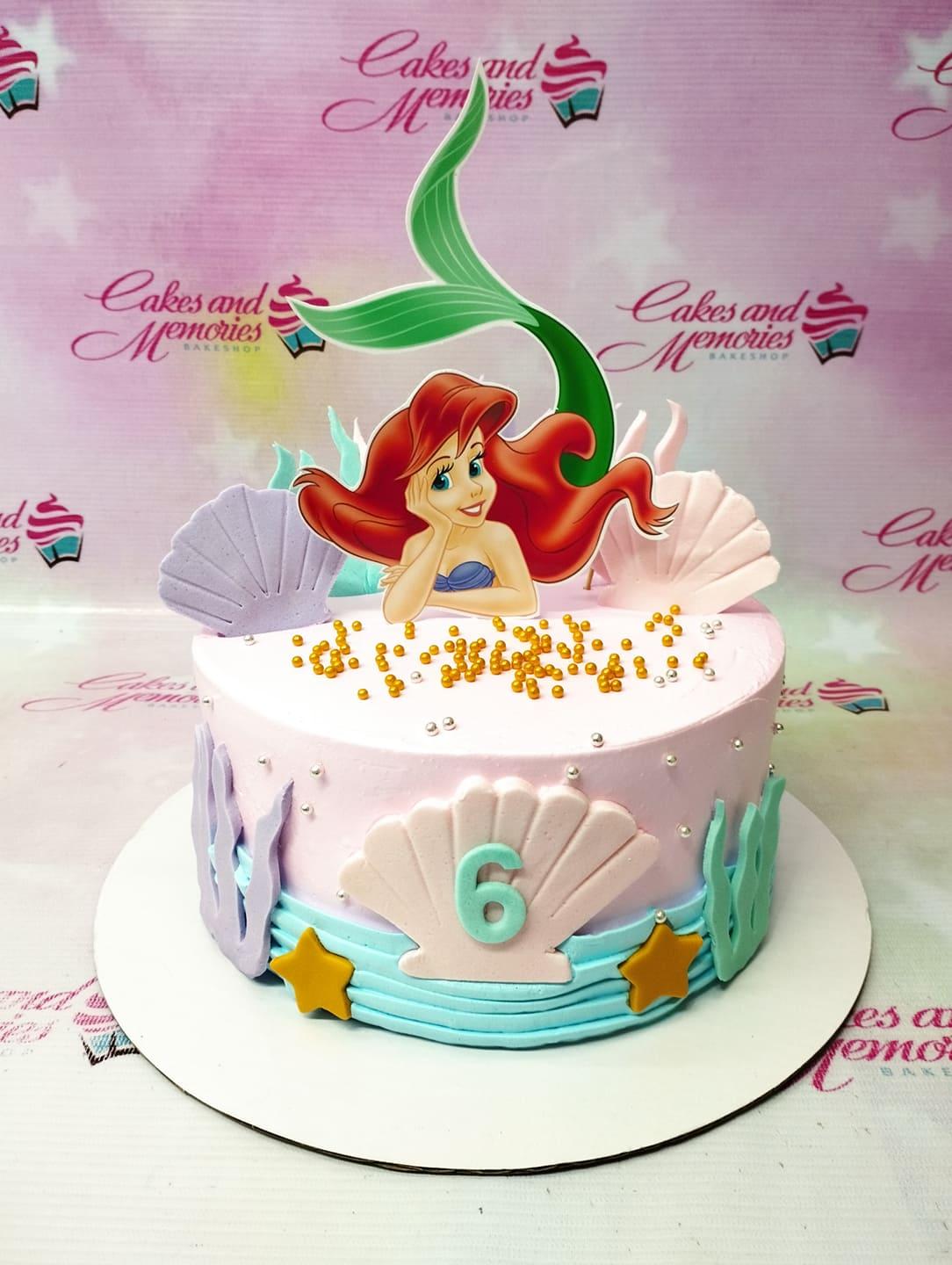 Pink Ariel The Little Mermaid birthday cake with purple and pink fondant shells, gold pearls, and a large mermaid tail topper.