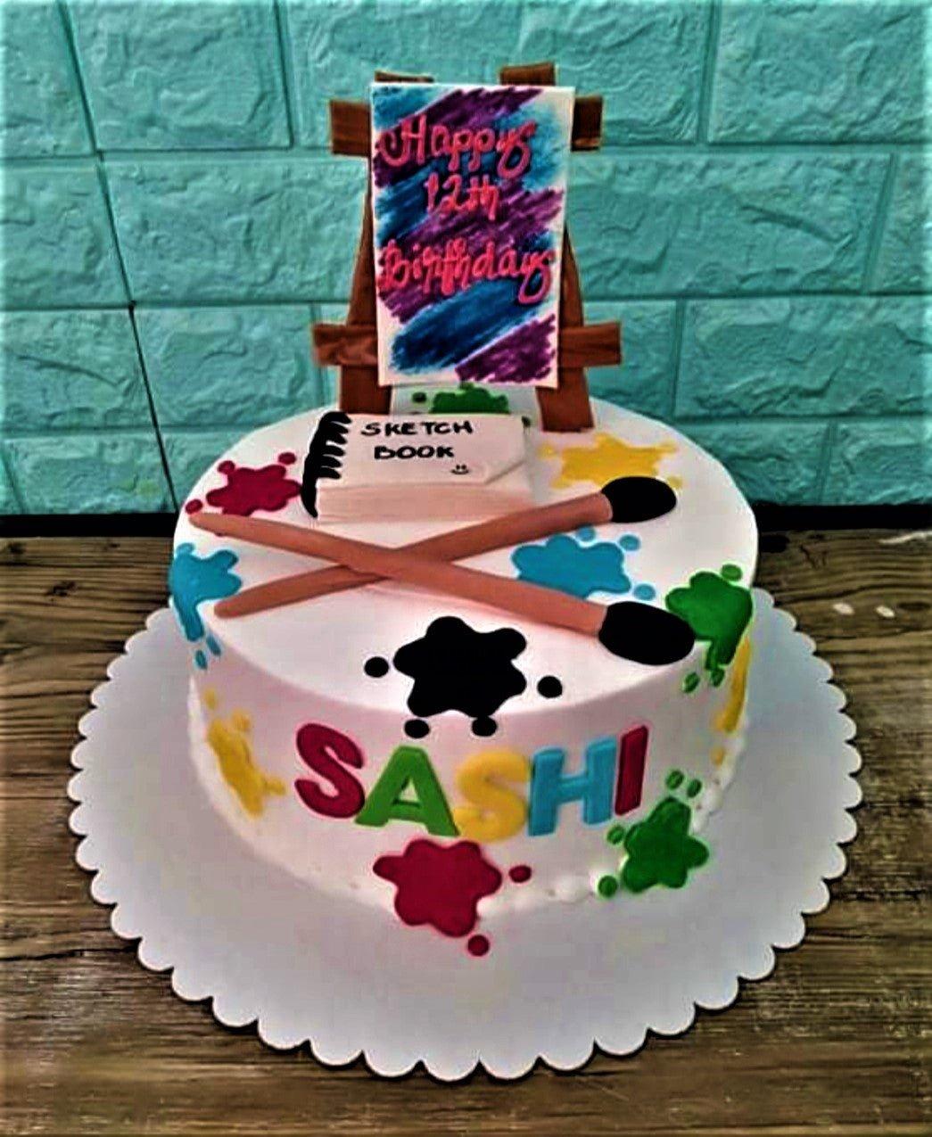 Artistic 12th birthday cake with an easel topper, sketch book, paint brushes, and colorful paint splatters on white icing.