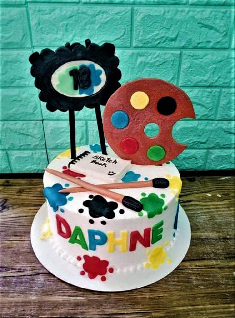 White 1-tier art themed birthday cake with colorful paint splatters, a fondant palette, sketch book, and paintbrushes.