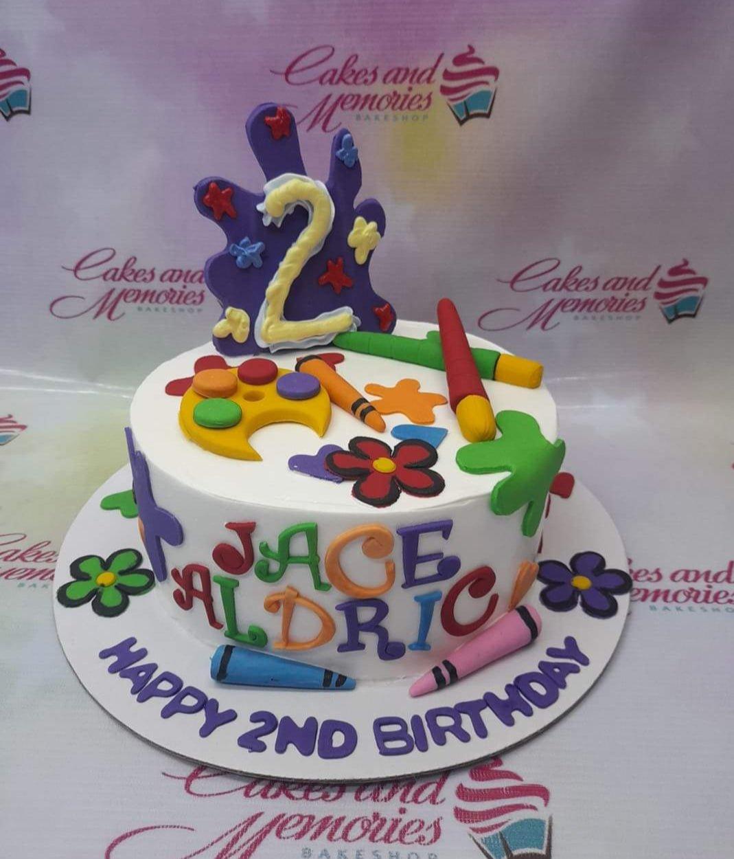 White round art-themed birthday cake with colorful fondant paint splats, crayons, and a paint palette.