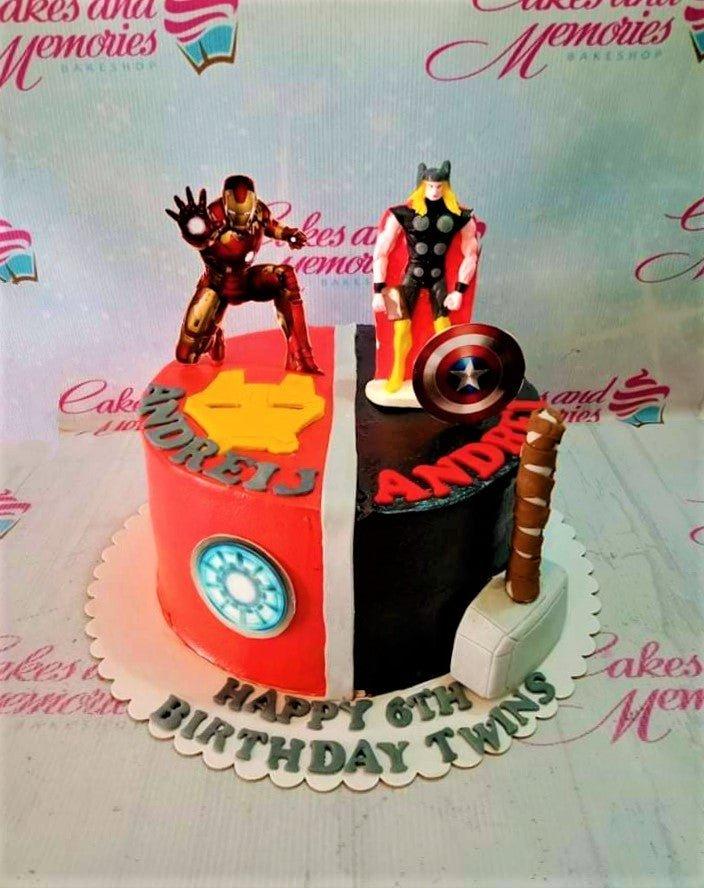 Avengers themed birthday cake for twins featuring Iron Man and Thor figurines with Captain America shield and Thor's hammer decorations.