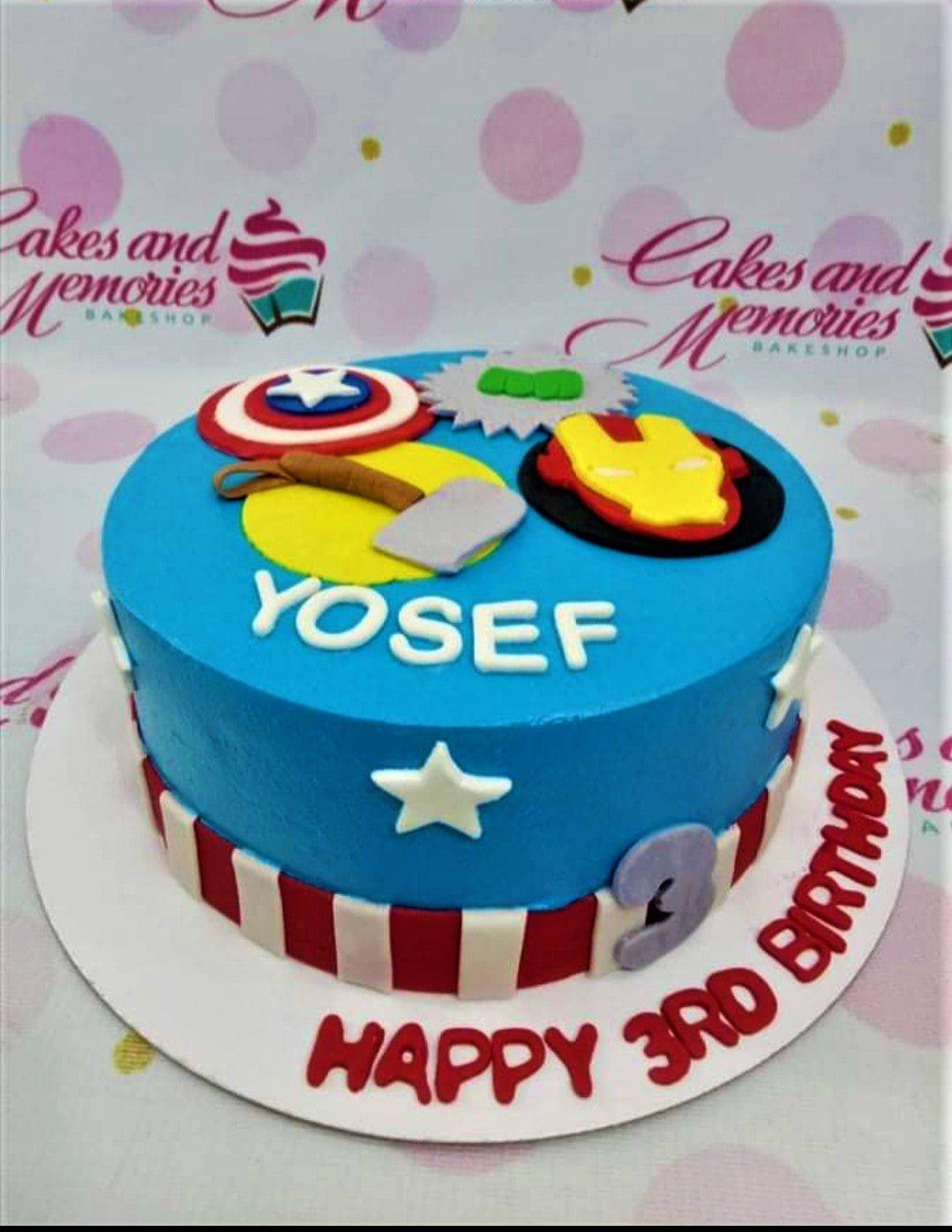 Blue Avengers themed birthday cake with Captain America shield, Thor's hammer, Hulk's fist, and Iron Man mask fondant toppers.