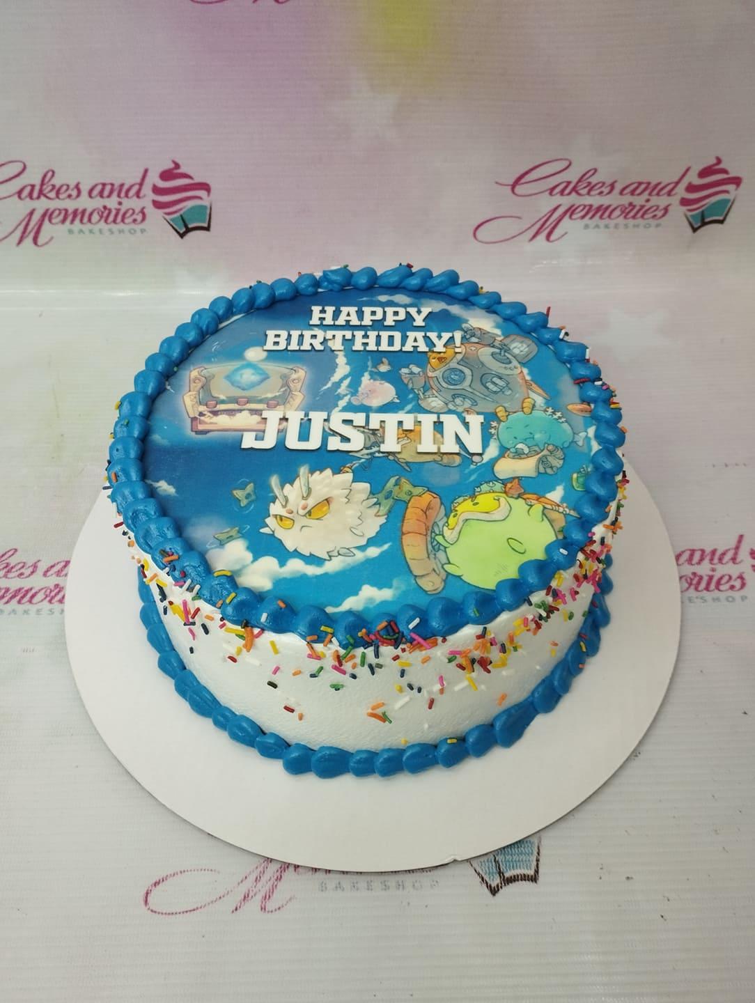 Round white cake with blue piped borders and a large Axie Infinity character printout on top, decorated with colorful sprinkles.