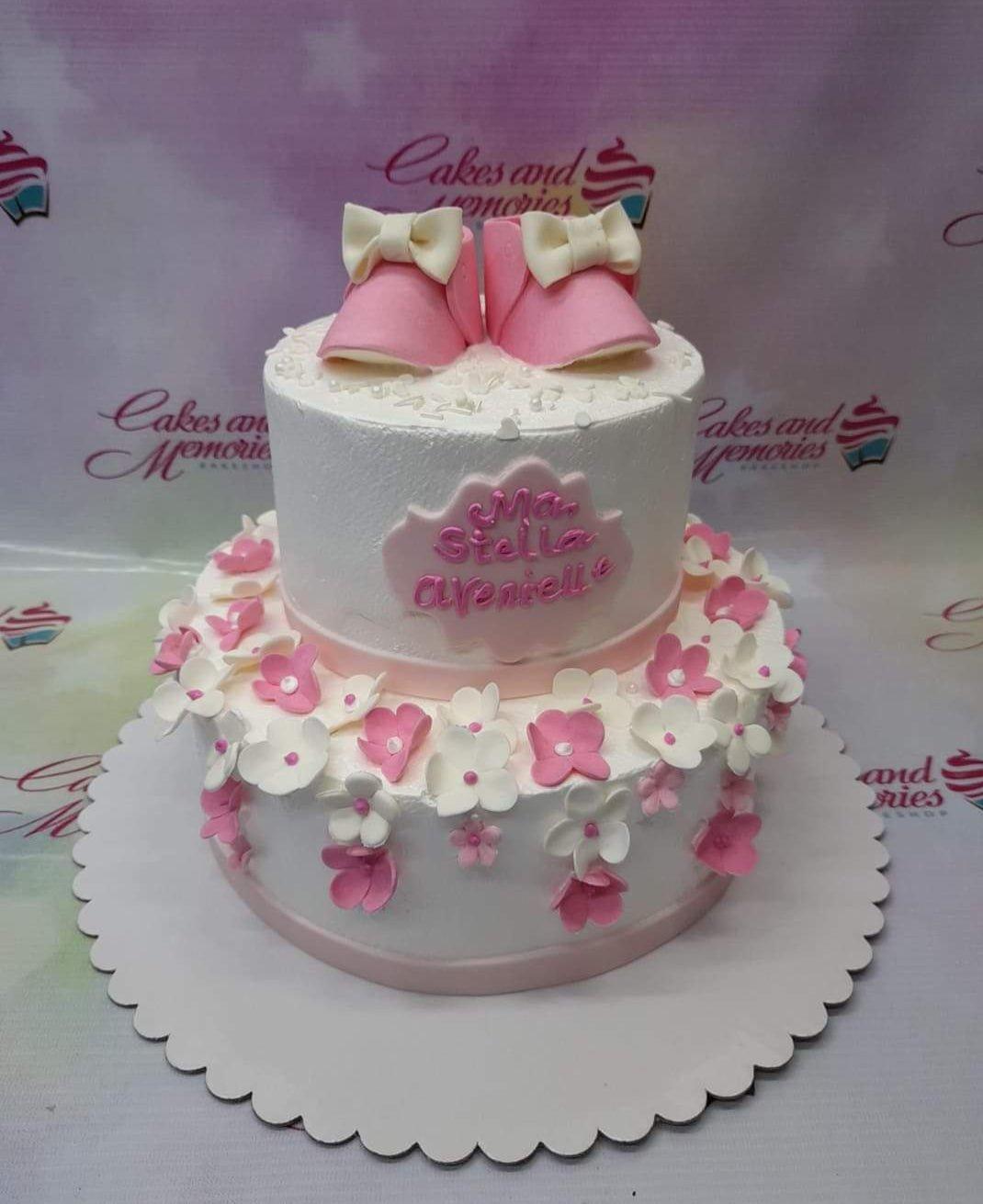 Two-tier white soft icing cake for a baby girl featuring pink fondant baby booties and edible flowers.