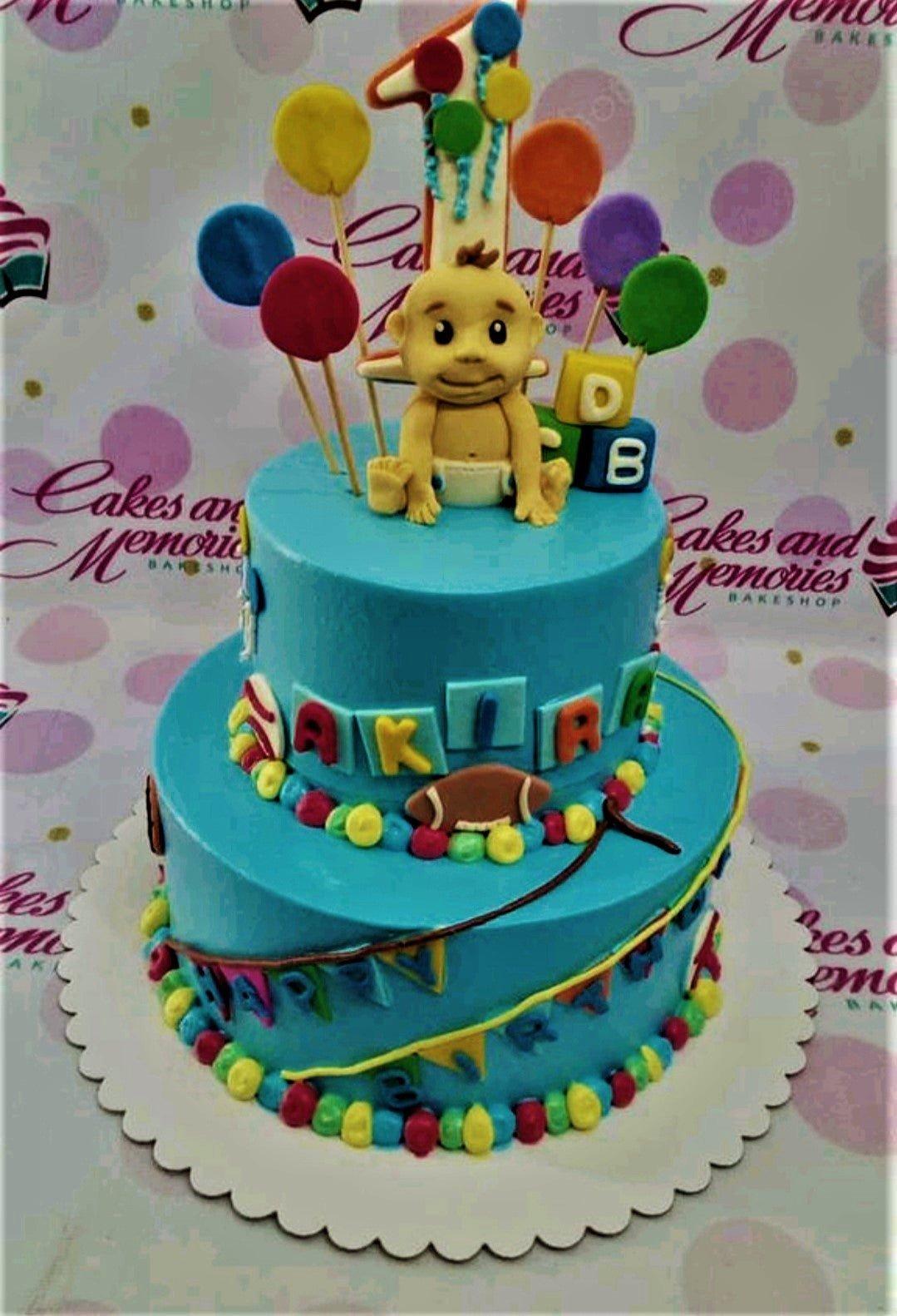 Blue 2-tier baby boy birthday cake with a fondant baby figure, colorful balloons, and alphabet blocks.