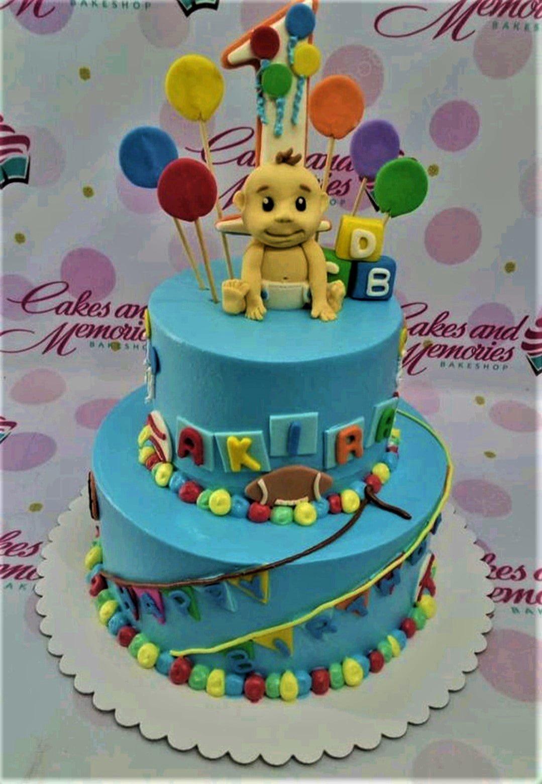 Two-tier blue fondant baby boy birthday cake with a fondant baby figure, colorful balloons, and alphabet blocks.