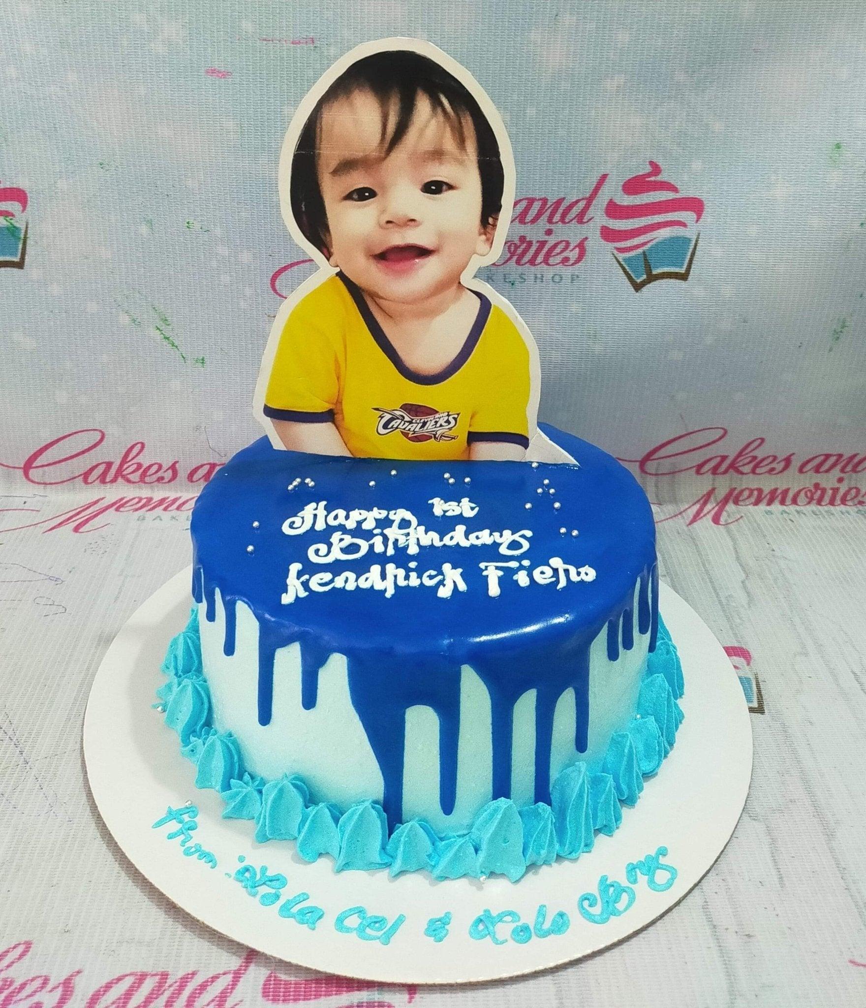 Light blue 1-tier birthday cake with dark blue drip, a large photo printout of a baby boy, and blue icing rosettes.