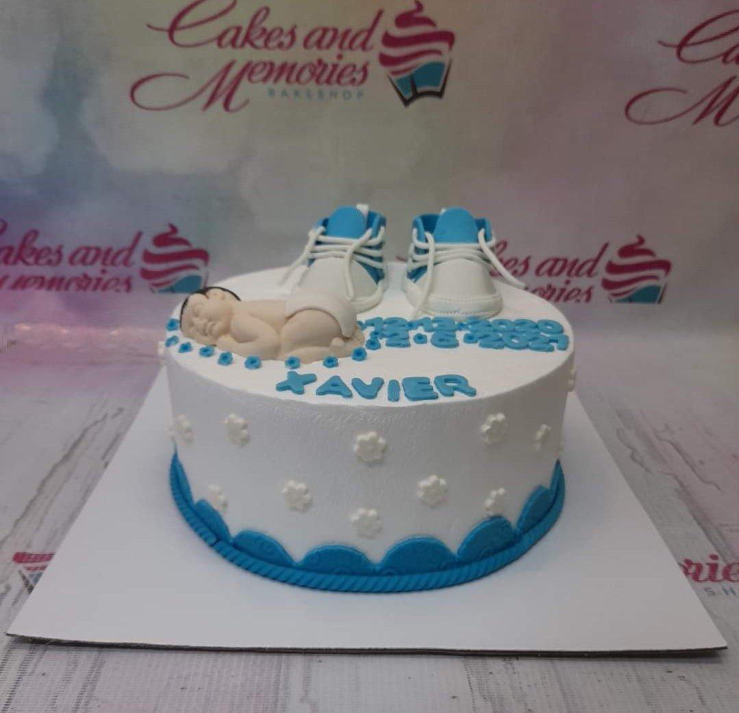 White 1-tier baby shower cake with blue accents, featuring a sleeping baby fondant figure and miniature blue and white sneakers.