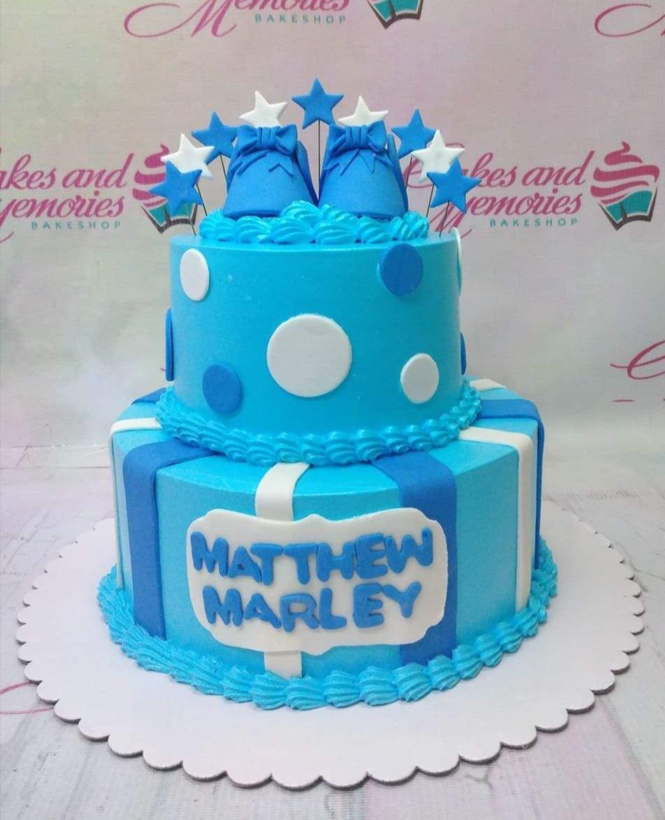 Two-tier blue and white polka dot and striped cake with fondant baby shoes and star toppers for a boy's birthday.