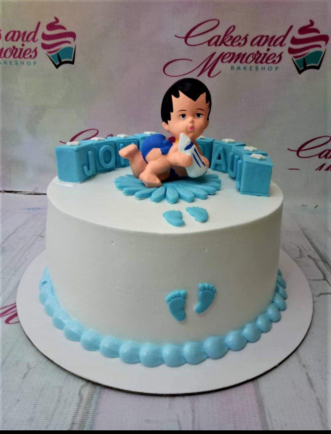 White round cake with a 3D baby figure topper holding a bottle, blue block letters spelling JONATHAN, and blue baby footprint accents.