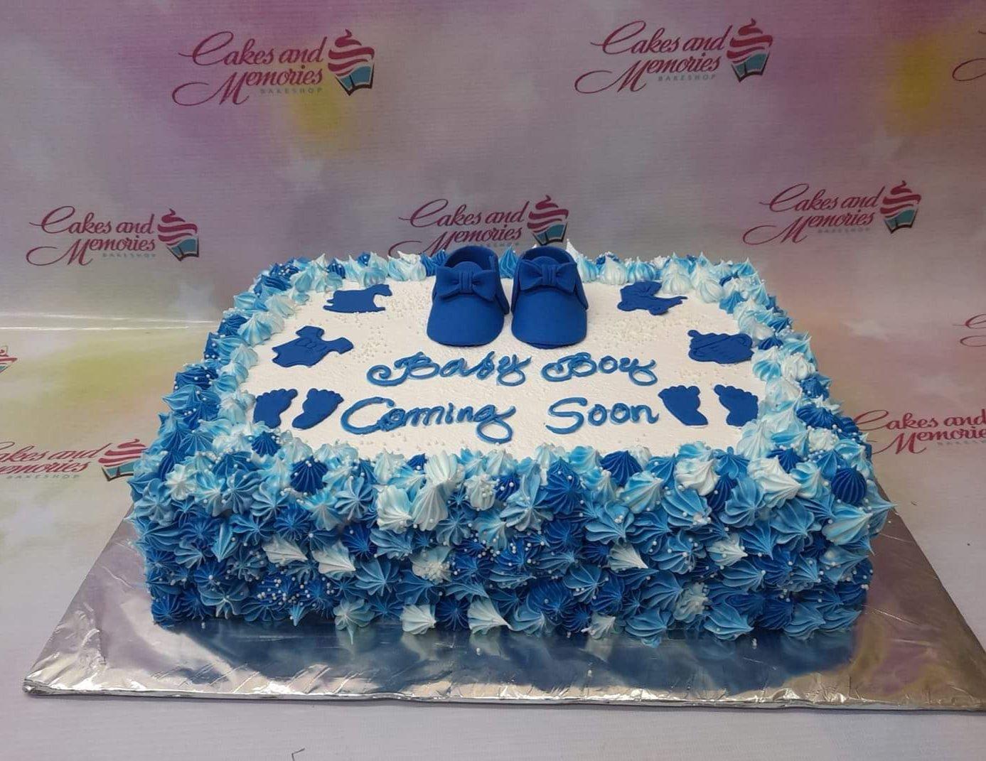Rectangle baby shower cake for a boy with blue and white icing rosettes, blue baby shoes, and baby-themed fondant cutouts.