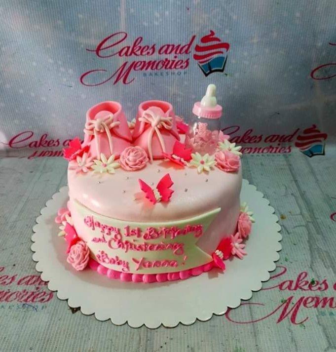 Pink baby girl christening cake with fondant baby shoes, a baby bottle, and pink butterflies on a light pink fondant base.