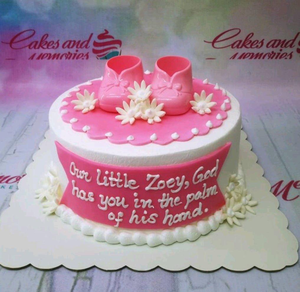 White soft-icing cake with pink baby shoes and white flowers, featuring a pink banner with a religious message.