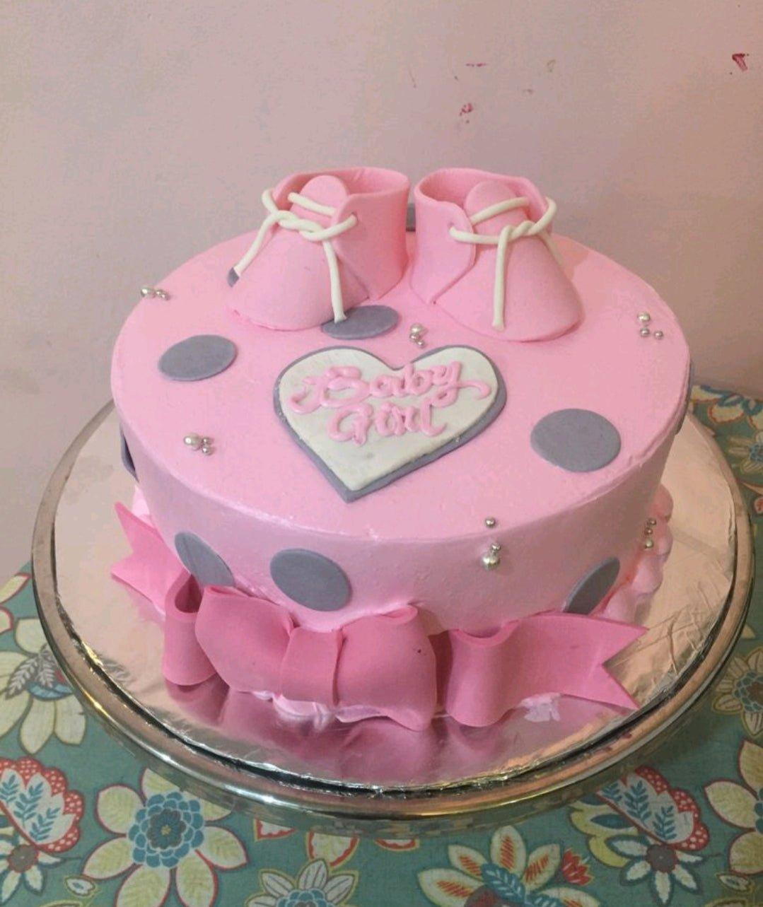 Pink baby girl cake with fondant baby shoes, grey polka dots, and a large pink bow at the base.