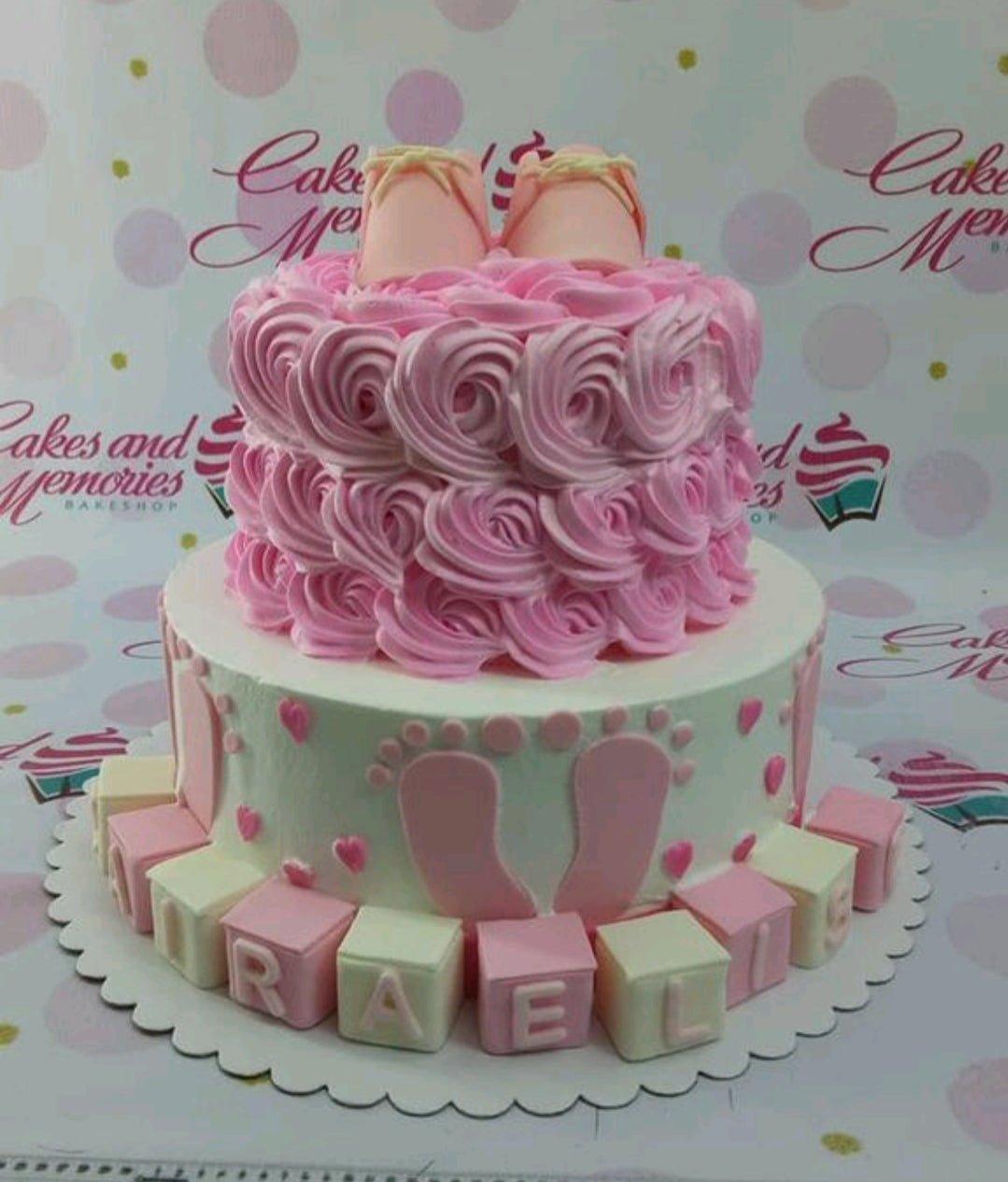 Two-tier pink and white baby girl cake with pink icing rosettes on the top tier and baby feet decorations on the bottom tier.