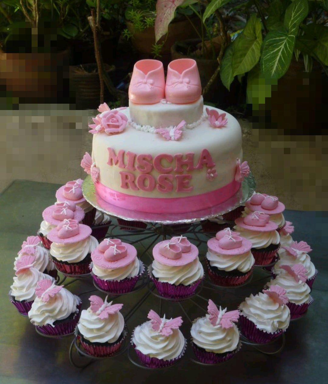 White fondant baby shower cake with pink baby shoes topper, pink butterflies, and matching cupcakes on a tiered stand.