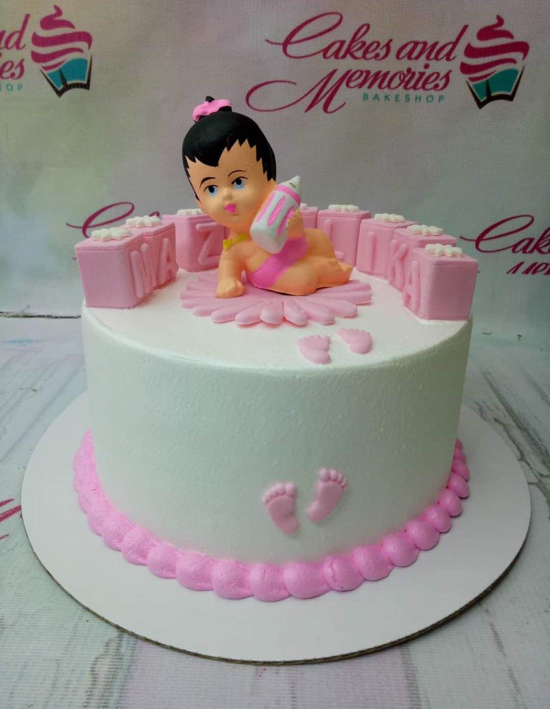 White soft-icing baby girl cake with a 3D fondant baby figure, pink blocks, and baby footprint decorations.