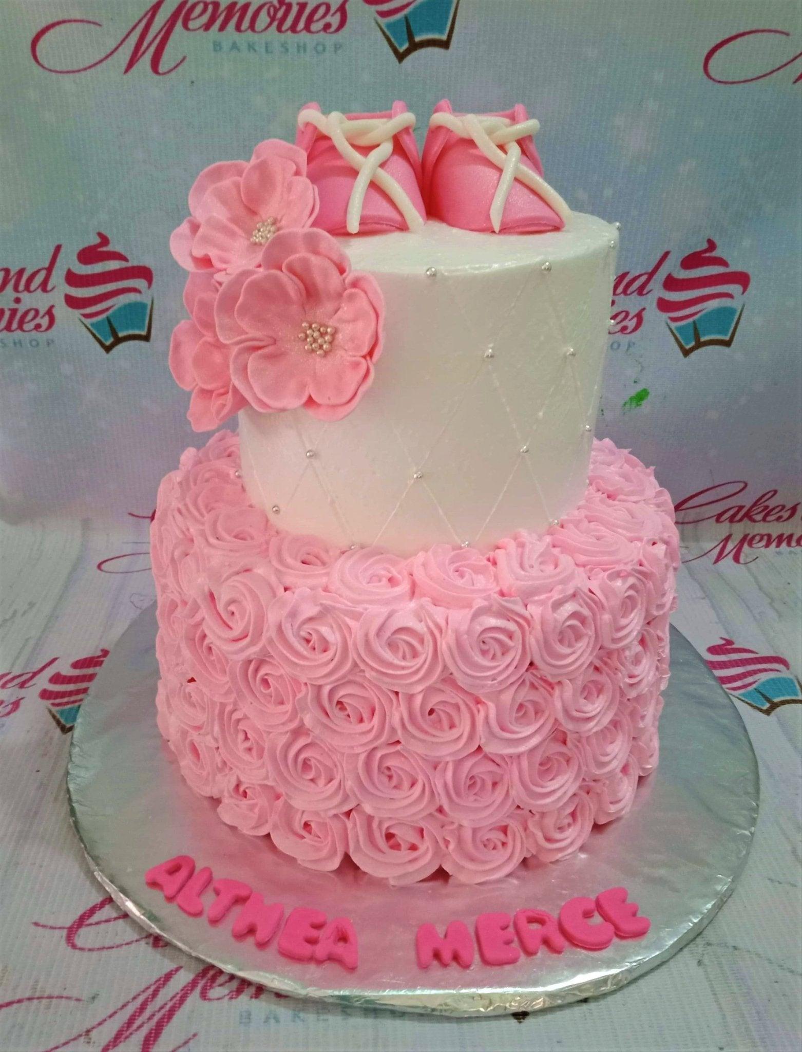 Two-tier pink and white baby shower cake with fondant baby shoes, pink icing rosettes, and quilted pattern with silver pearls.