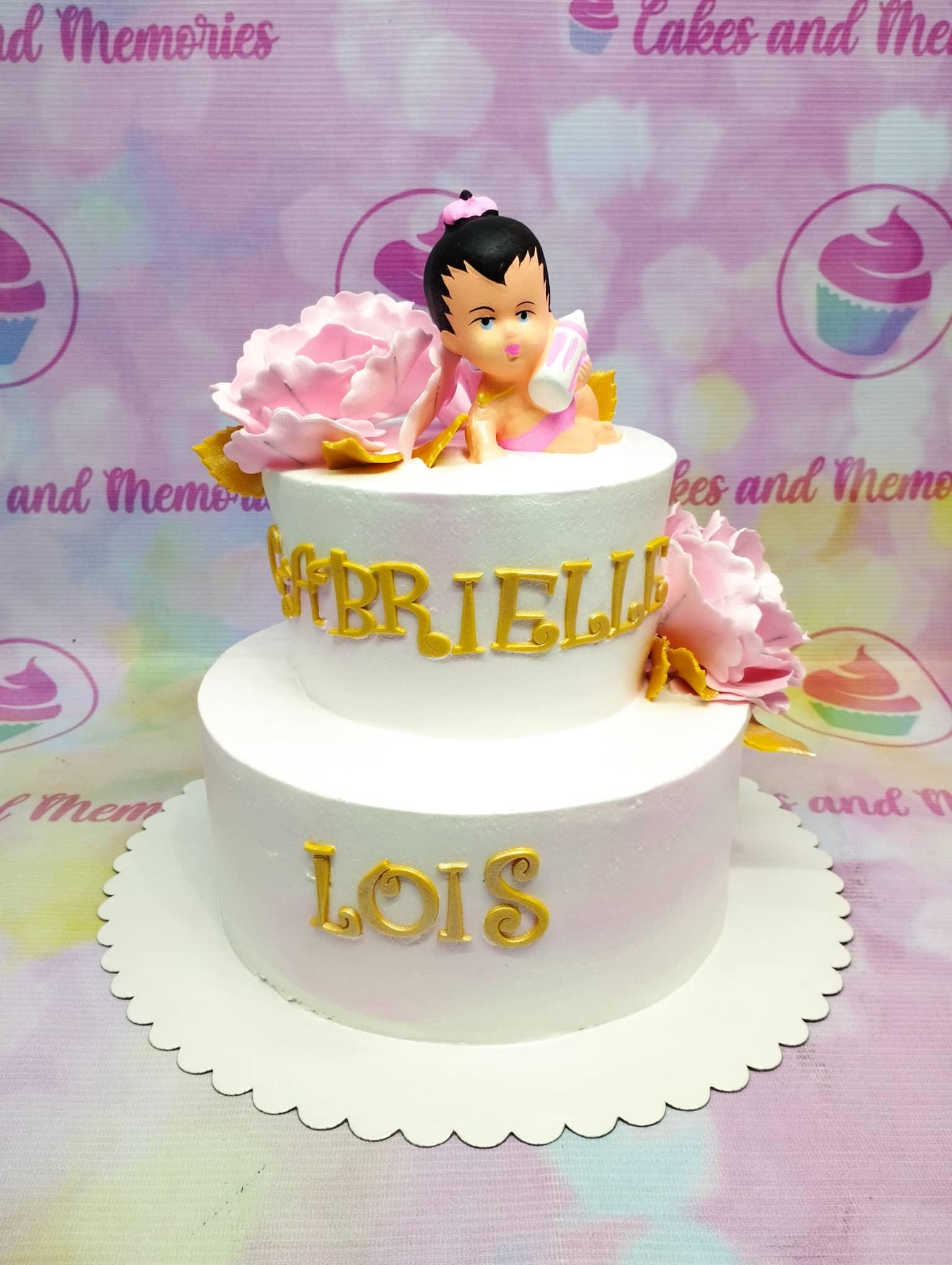 White 2-tier baby girl birthday cake with pink fondant flowers and a baby figurine topper holding a bottle.