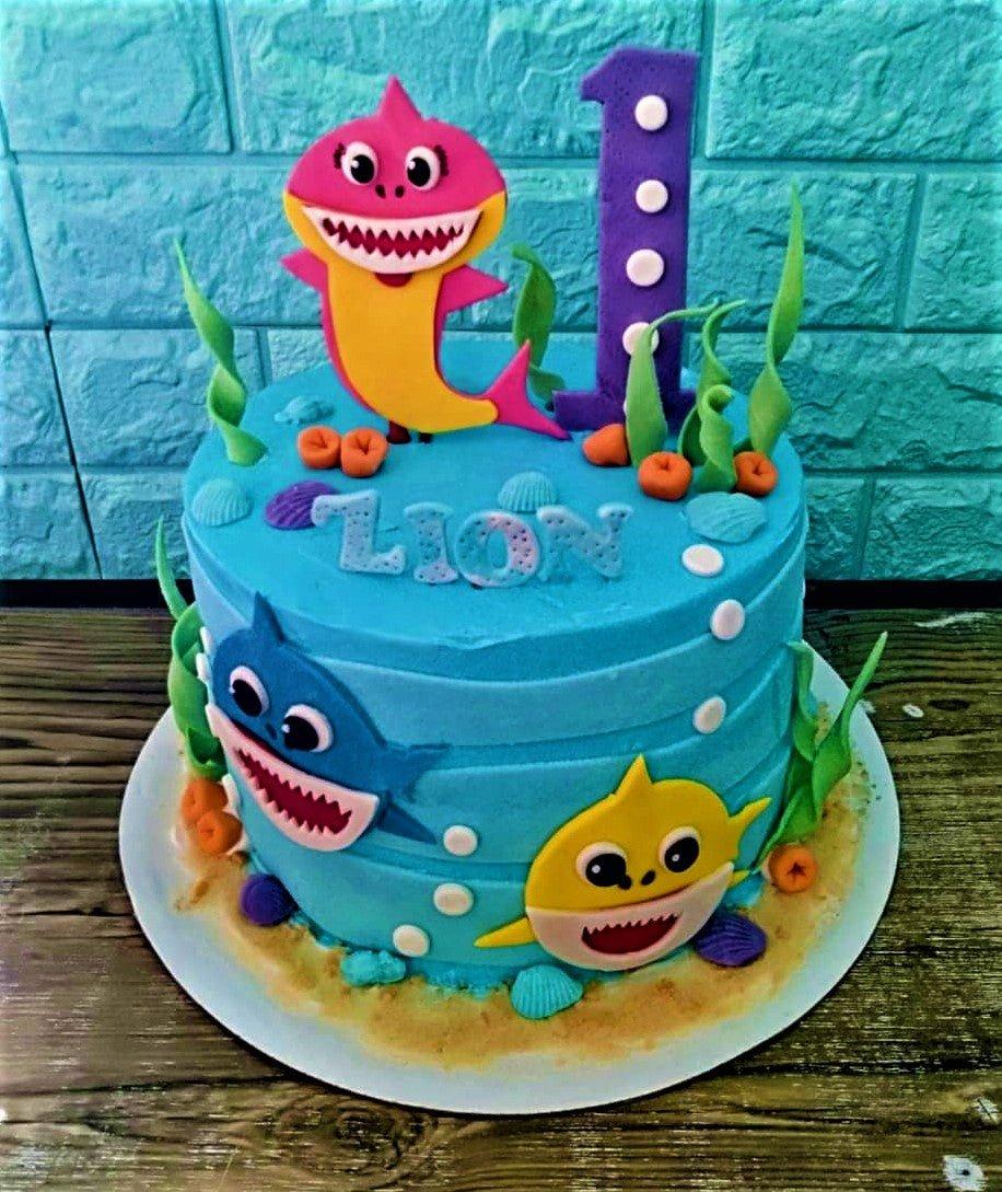 Blue Baby Shark themed birthday cake with pink, blue, and yellow shark characters, purple number one, and underwater fondant decorations.