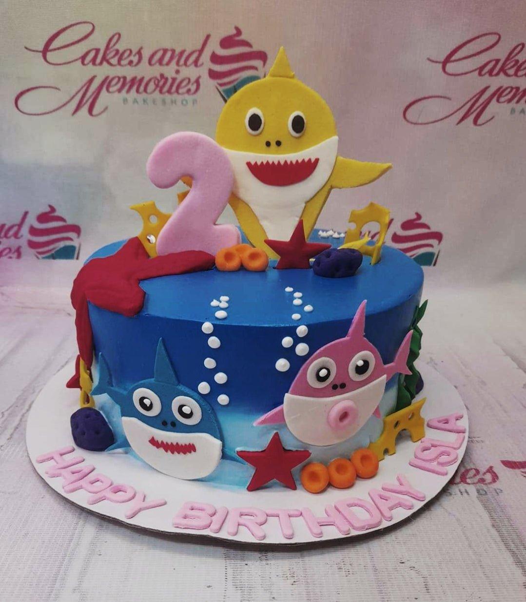 Blue Baby Shark themed birthday cake with fondant shark figures, coral, and bubbles on a white base board.