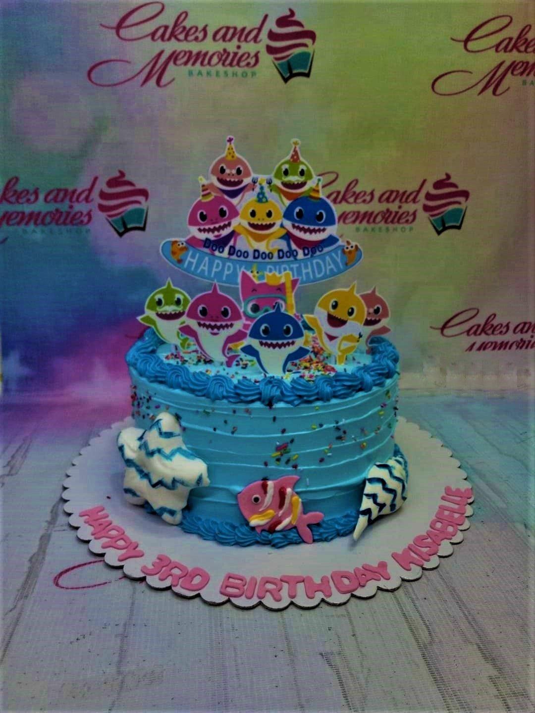 Blue Baby Shark themed birthday cake with colorful character printouts, icing rosettes, and sea-themed fondant decorations.