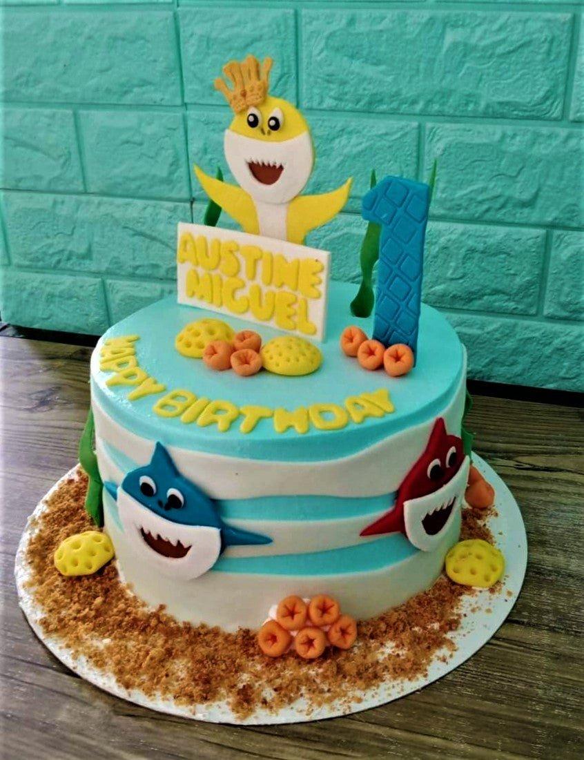 Baby Shark themed blue and white striped cake with fondant shark figures and coral decorations for a first birthday.