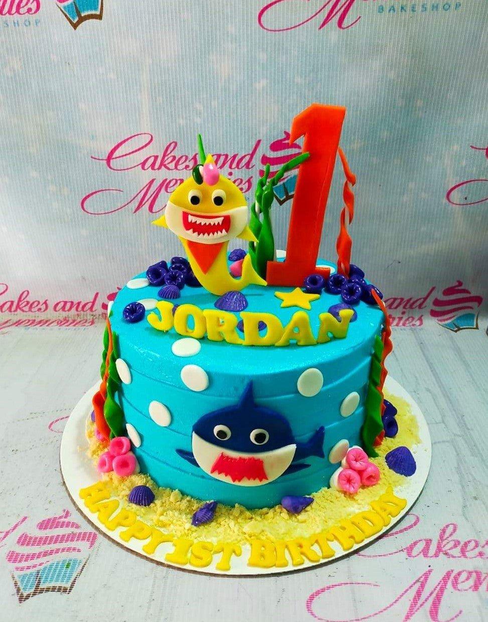 Blue Baby Shark themed birthday cake with yellow and blue shark toppers, seaweed, and coral decorations.