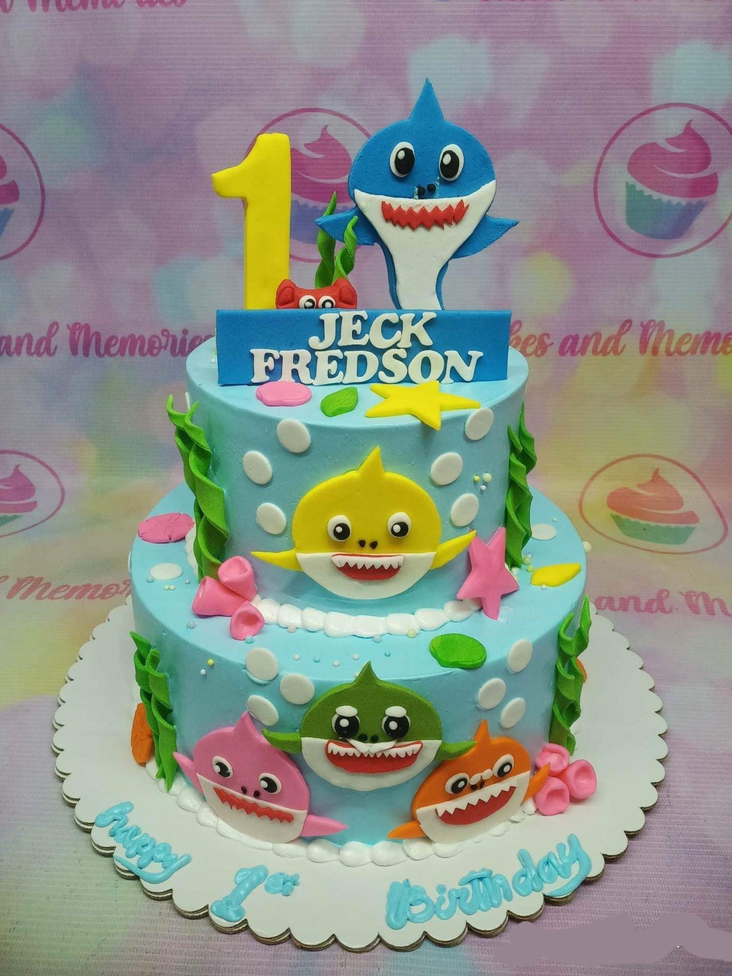 Two-tier light blue Baby Shark themed birthday cake with colorful fondant shark characters, seaweed, and a yellow number one topper.