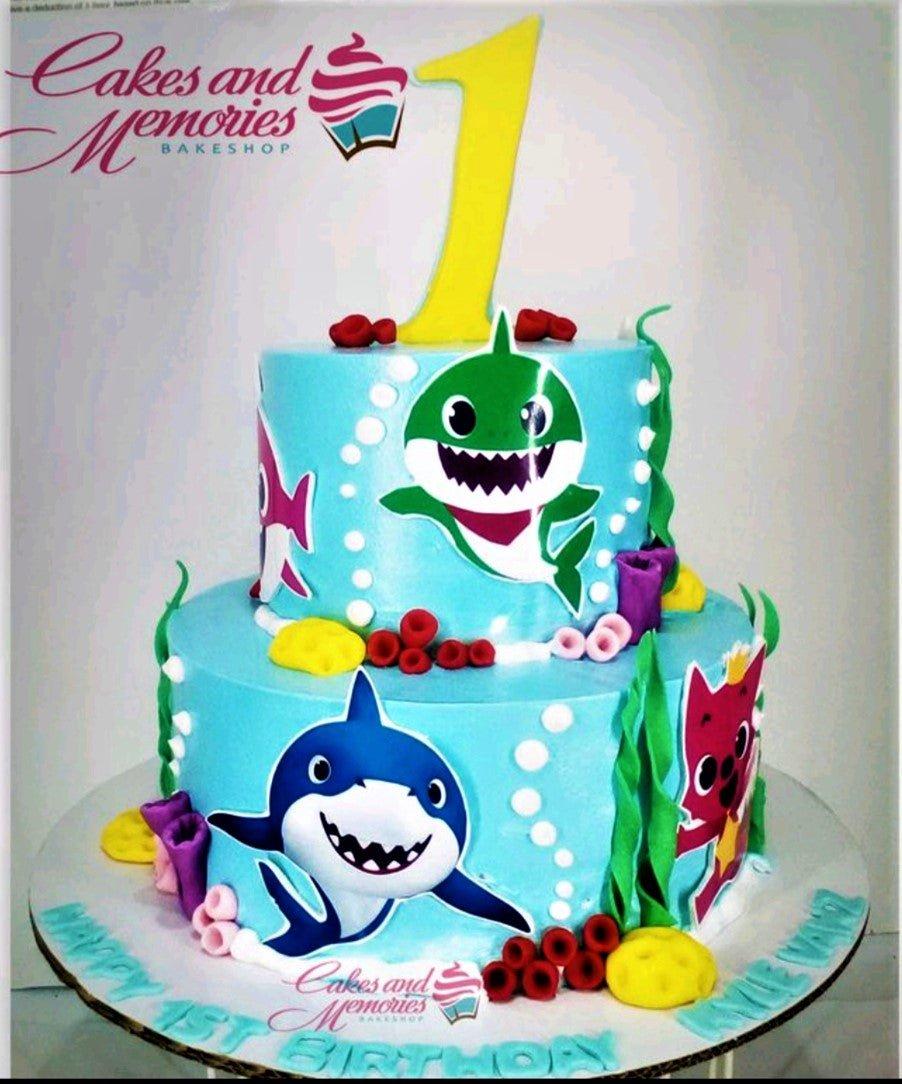 Two-tier light blue Baby Shark themed birthday cake with colorful underwater fondant decorations and character printouts.