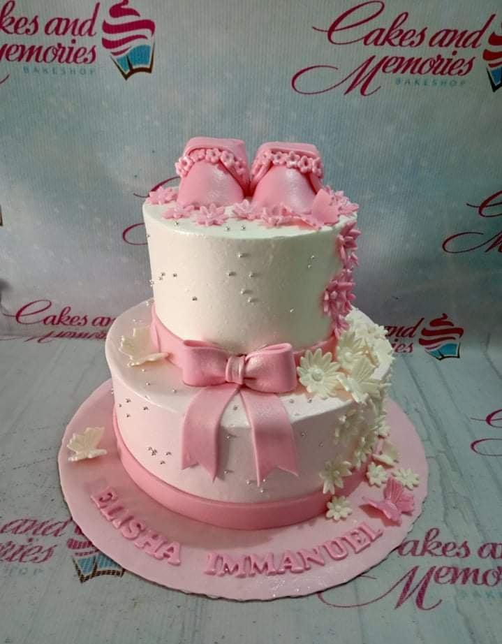 Two-tier pink and white baby shower cake with fondant baby shoes, pink bow, and white and pink flowers.
