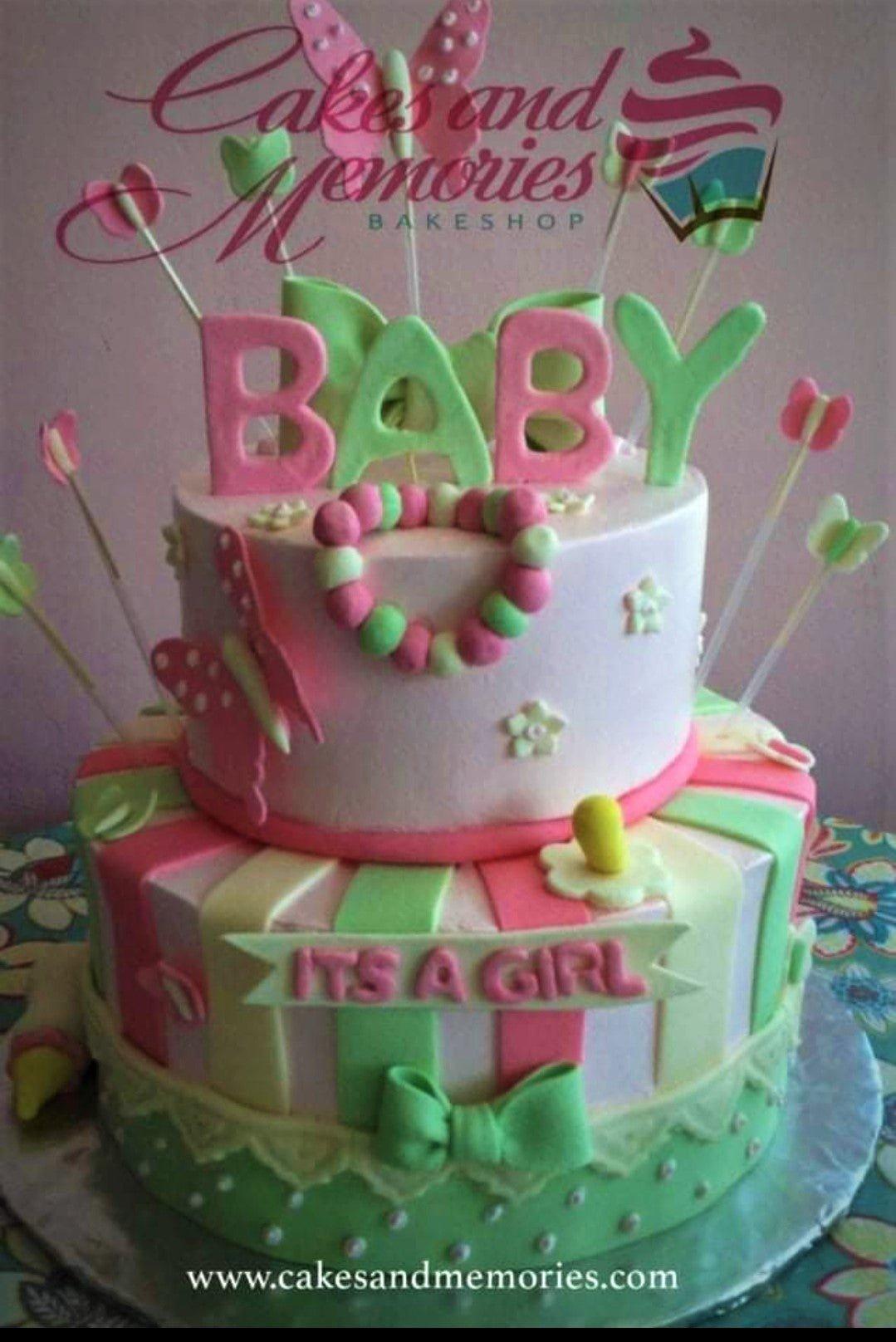 Pink and green 2-tier baby shower cake with fondant butterflies, a baby rattle, and 'ITS A GIRL' message.