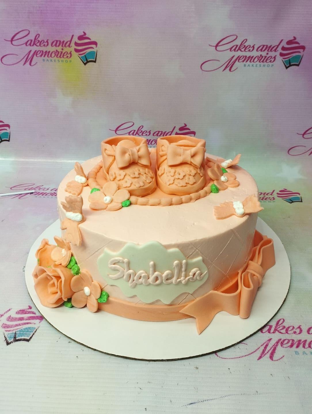 Peach-colored baby shower cake with fondant baby booties, flowers, and a large bow on the side.
